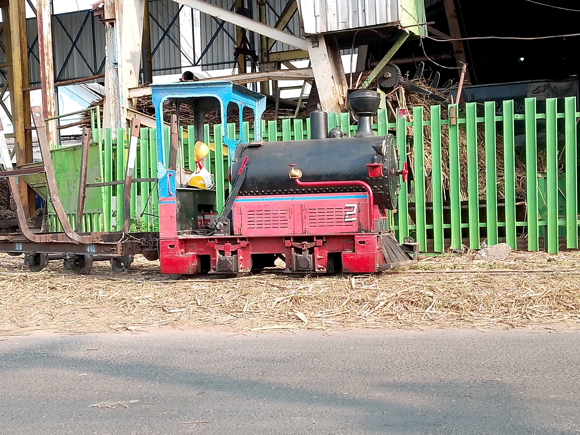 A miniature train, painted red and blue with a black engine, sits on a bed of dry grass in front of a bright green metal fence. The train has a visible number "2" on its side. Behind the fence, a large structure with metal beams and corrugated roofing appears to be an industrial building or warehouse. The ground in front of the train is paved with asphalt, suggesting a road or walkway. The scene is lit by natural daylight, casting shadows on the asphalt. The background is partially obscured by the train and fence, but hints at a rural or industrial setting. The image is taken from a slightly low angle, looking towards the train and its surroundings.