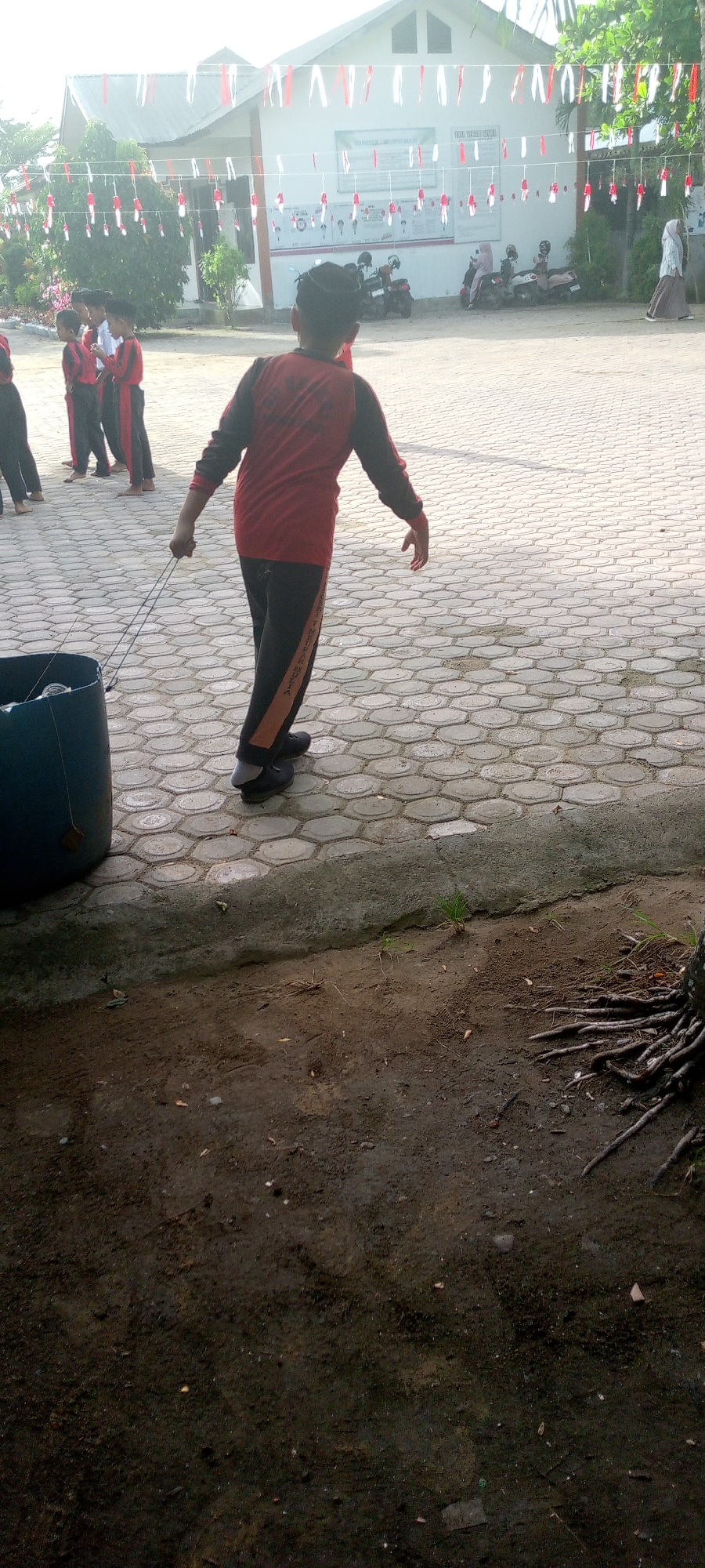 In the city of Baratpayaitek, Indonesia, an image captures a scene in an outdoor area, possibly a schoolyard, during daylight. In the foreground, a young person, viewed from behind, is wearing a red long-sleeved shirt with black sleeves and black pants with an orange stripe on the side. They are pulling a large, dark blue cylindrical object with two ropes. The ground is paved with hexagonal gray tiles, and a patch of dirt with exposed tree roots is visible in the lower portion of the image. In the background, a building with white walls is decorated with red and white banners and streamers, suggesting a celebration or event. Several children in similar uniforms are standing near the building, and some motorcycles are parked in front of it. A woman in a headscarf is walking away from the building on the right side of the frame. The overall lighting indicates a sunny day.
