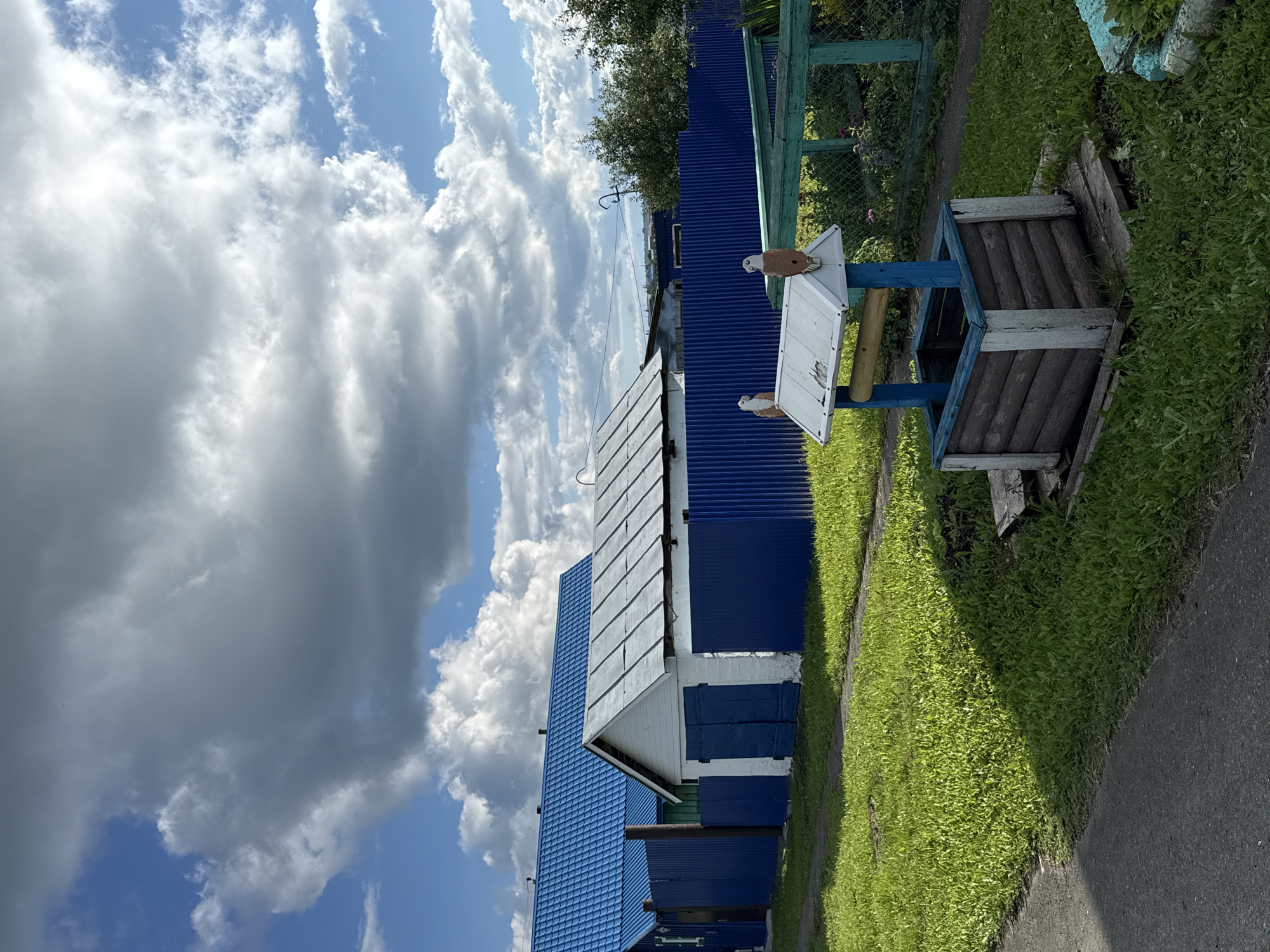 The image depicts a rustic scene in the village of Bolshetyurish, Russia, under a partly cloudy sky. In the foreground, a wooden well with a blue and white trim stands on a wooden platform surrounded by green grass. Two wooden bird figures are perched on the well's roof. A blue corrugated metal fence runs across the midground, separating the yard from a grassy area and a paved path. Behind the fence, a white building with blue doors and a silver roof is visible, alongside another building with a bright blue roof. Lush green grass dominates the ground, with some shadows suggesting the presence of trees or structures casting them. The overall atmosphere is one of rural simplicity.