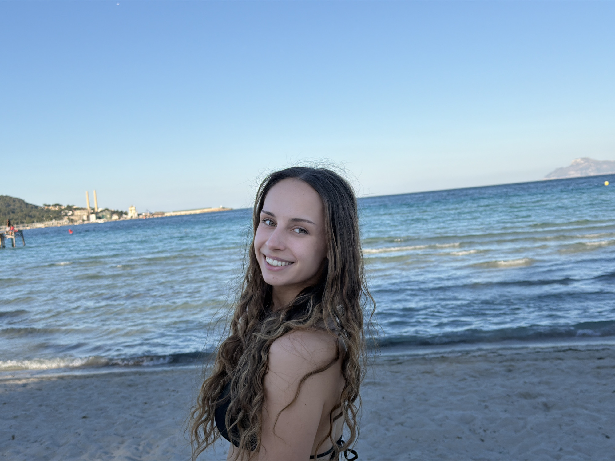 A young woman with long, curly brown hair stands on a sandy beach, smiling and looking over her shoulder at the camera. She is wearing a black bikini top. Behind her is the Mediterranean Sea, with gentle waves rolling onto the shore. In the distance, buildings are visible along the coastline of Alcúdia, Spain, and a mountainous area can be seen on the horizon. A pier structure is visible to the left. The sky is clear and blue. The sand appears to be light in color, and the water is a mixture of blue and turquoise hues.