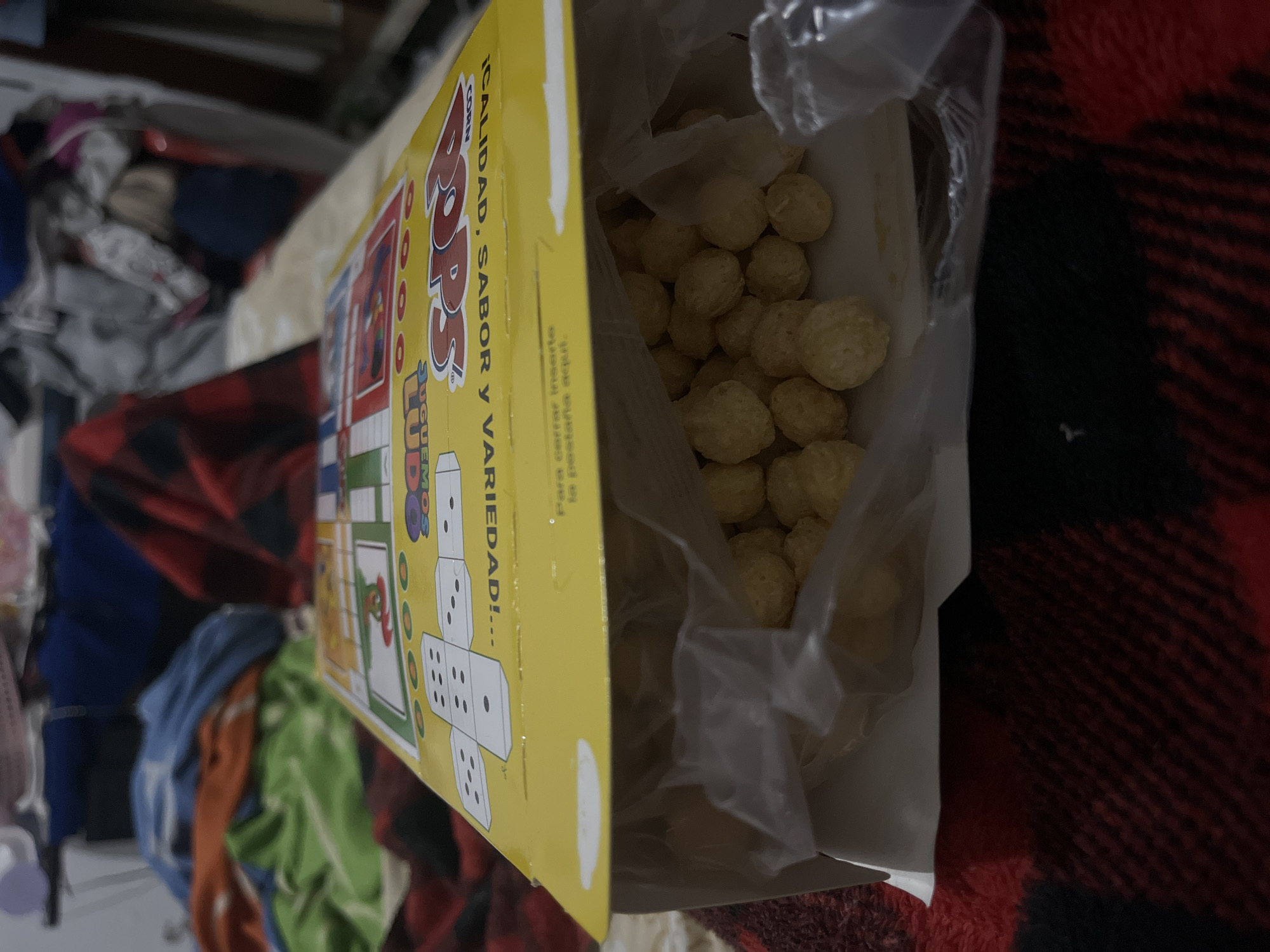 The image shows an open box of "Corn Pops" brand cereal, with a plastic bag containing the cereal inside the box. The box is yellow with red lettering. The cereal consists of small, round, yellow puffs. The box is partially resting on a red and black checkered blanket. In the background, there is a cluttered area with various clothing items. There is no indication of location within the city of Santa Ana, Venezuela.