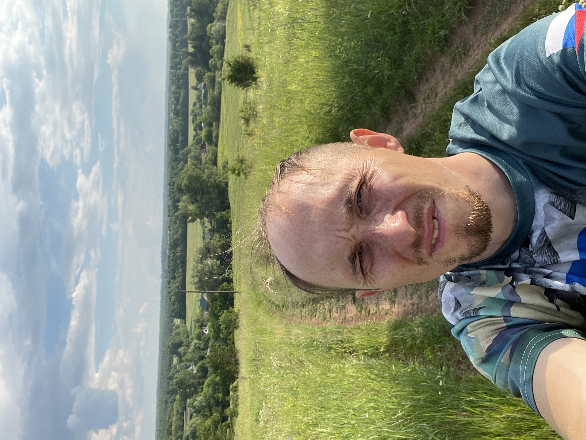 A selfie is taken by a man in his early 30s with light skin, receding hairline, and a short beard. He is wearing a camouflage t-shirt and standing on a dirt path in a grassy field. In the background, there are trees, houses, and a blue sky with clouds. The scene appears to be in the village of Nizhnii Bugrysh, Russia.