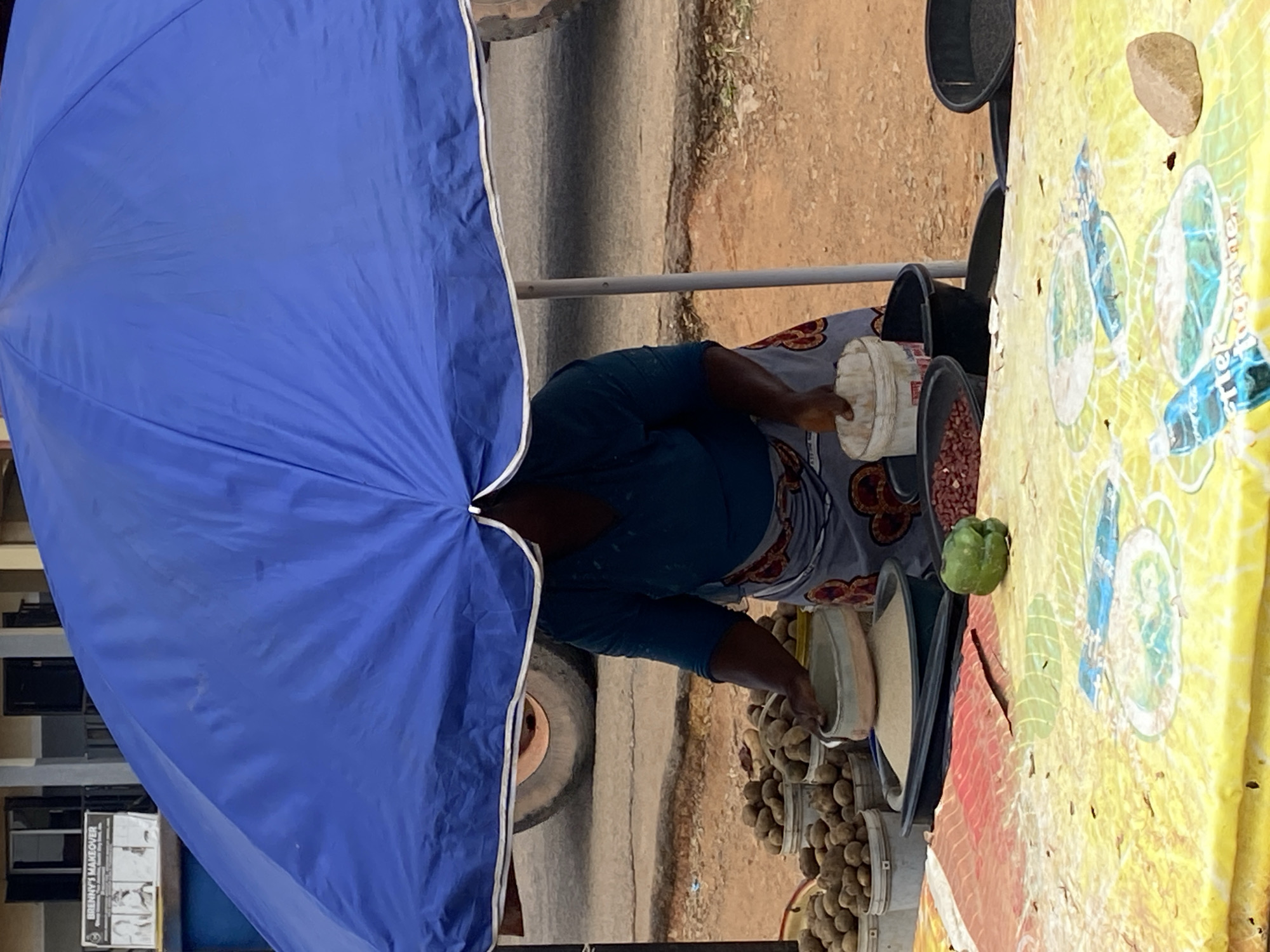 In Gwong, Nigeria, a vendor stands under a large blue umbrella. She is wearing a teal shirt and a patterned skirt. In front of her, on a table covered with a yellow and colorful patterned cloth, are several buckets and bowls containing produce and grains. To her left, there are buckets filled with potatoes. In the center, she is scooping sand or grain from a light grey bowl into a black container. To her right, there are bowls of red beans and another empty black container. A green bell pepper sits on the table to her left. In the background, a large tire from a vehicle is visible. A sign with "BRENNY'S MAKEOVER" is posted on a building behind the umbrella. The ground is a dirt and gravel surface.