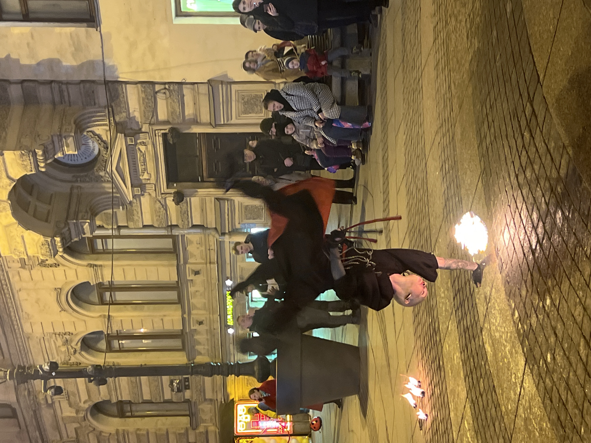 The image shows a street performer in the city of Saint Petersburg, Russia. He is balanced on one hand while holding a flaming torch in his other hand. The performer is wearing black clothing with a red cape. A crowd of people is watching the performance. The performance is occurring on a street paved with cobblestones. In the background, there are buildings with architectural details and windows. A streetlight is on the left of the frame. A glowing advertisement is visible at the bottom.