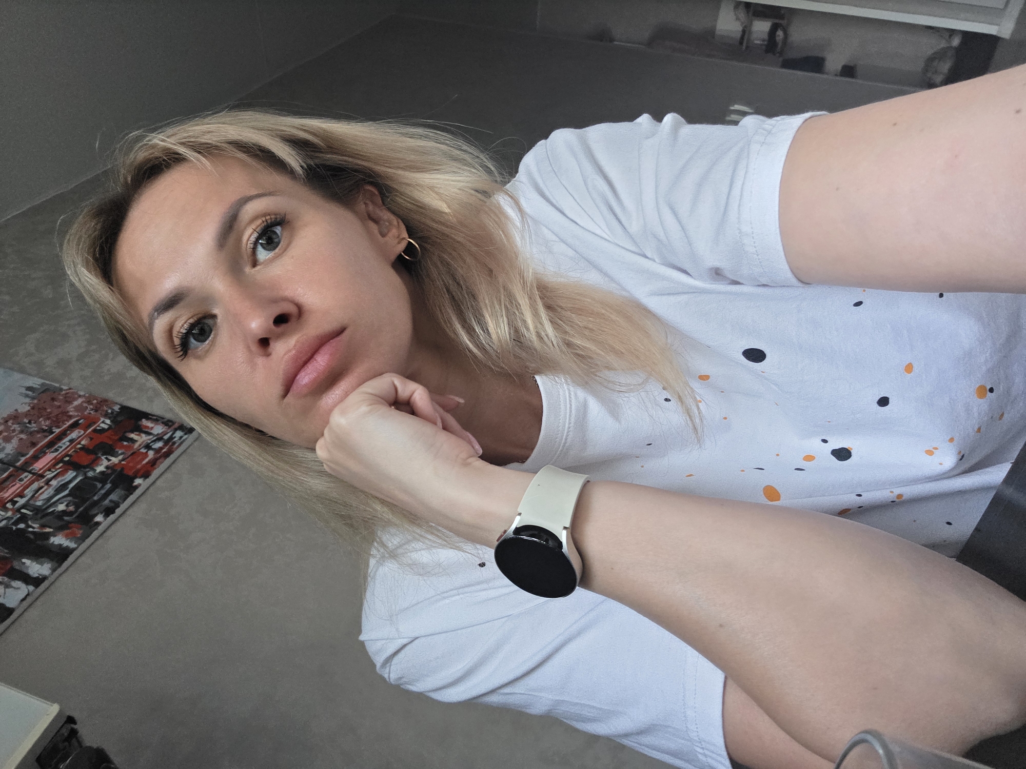 A woman with blonde hair is posing indoors for a selfie. Her head is tilted, and her hand is supporting her chin. She is wearing a white t-shirt with black and orange splatter patterns, a gold hoop earring, and a smartwatch with a light-colored band. A painting is visible on the wall behind her, along with other furniture or shelves in the background. The location is unknown and cannot be confidently determined as Chaikovsky, Russia.