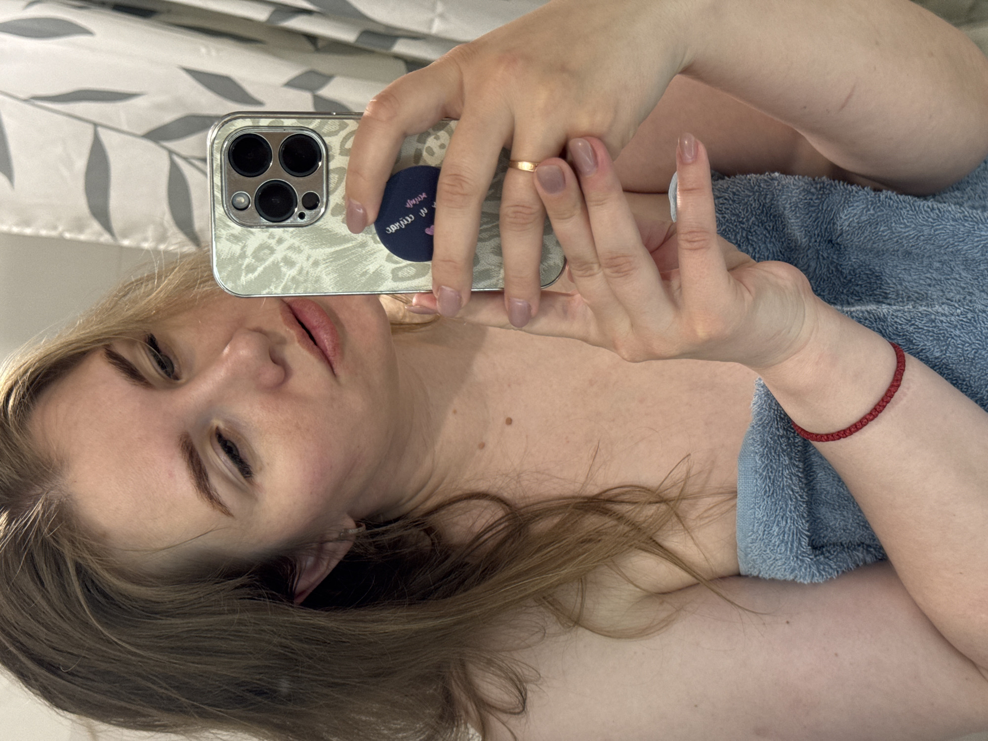 The image depicts a person holding a smartphone, presumably taking a selfie. The phone is an iPhone with multiple rear cameras and a patterned case. The person is wearing a gold ring on their ring finger and a red bracelet on their left wrist. They appear to be partially covered by a blue towel. The background suggests a bathroom setting with a white and gray patterned shower curtain. The location in the city of Ферма, Russia is not discernible from the image.