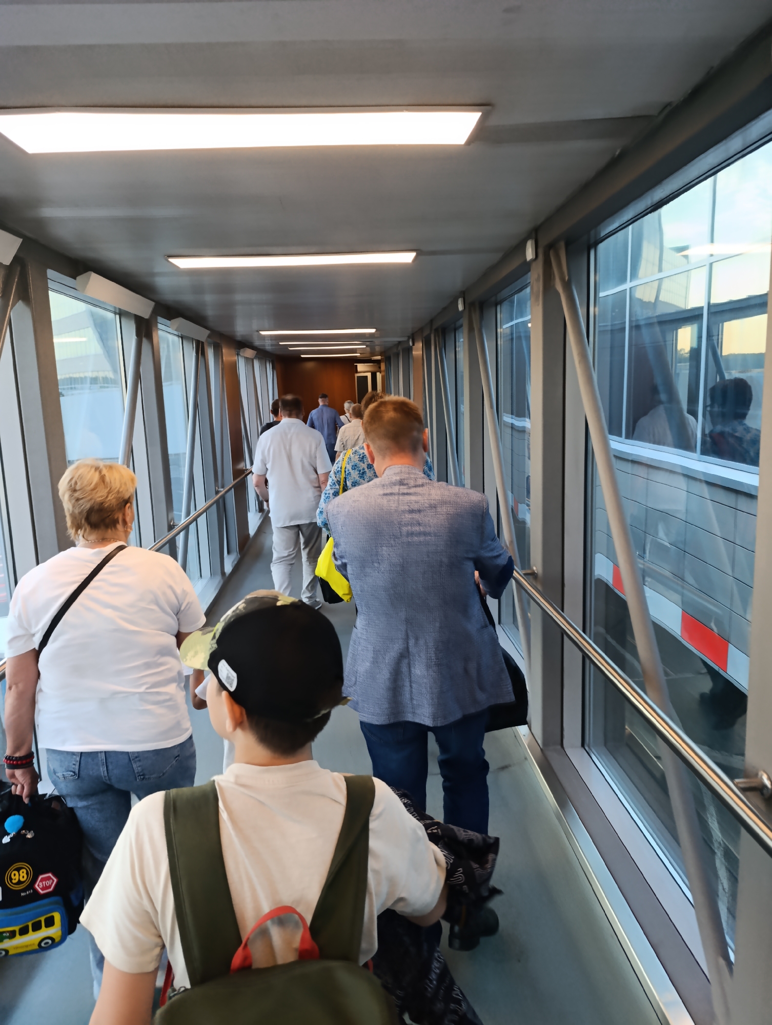 In Domodedovo, Russia, passengers are walking down an airport jet bridge. The jet bridge has large windows on the right side, offering a view of the tarmac and buildings outside. Overhead, fluorescent lights illuminate the walkway. Several people are visible, walking in the same direction. In the foreground, a young boy with a backpack and a baseball cap is walking away from the camera. To his left, a woman in a white t-shirt and jeans is also walking forward. Further ahead, a man in a blue suit is walking with a group of other passengers. A woman with a yellow bag is also in the group. The overall scene depicts the movement of people through an airport terminal.