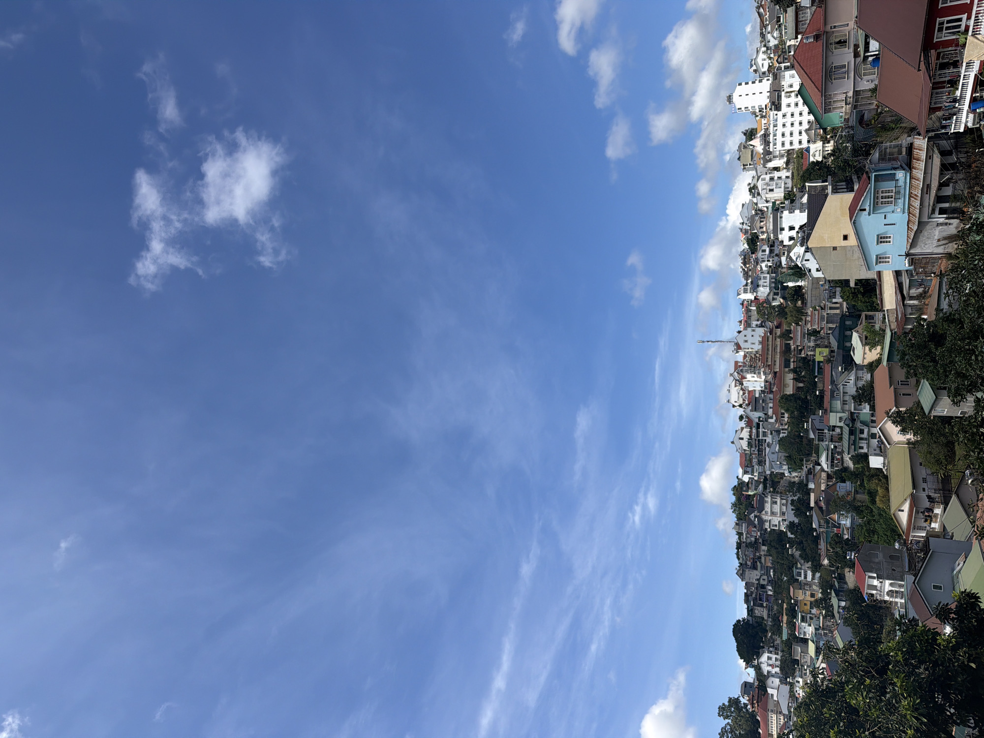 The image shows a dense hillside residential area in Da Lat, Vietnam, under a bright blue sky with scattered white clouds. The buildings are of various sizes and architectural styles, many with pitched roofs in red, green, and brown. The houses are clustered closely together, cascading down the slope, with some surrounded by lush green trees and foliage. The overall impression is one of a vibrant, somewhat chaotic urban landscape integrated with nature.