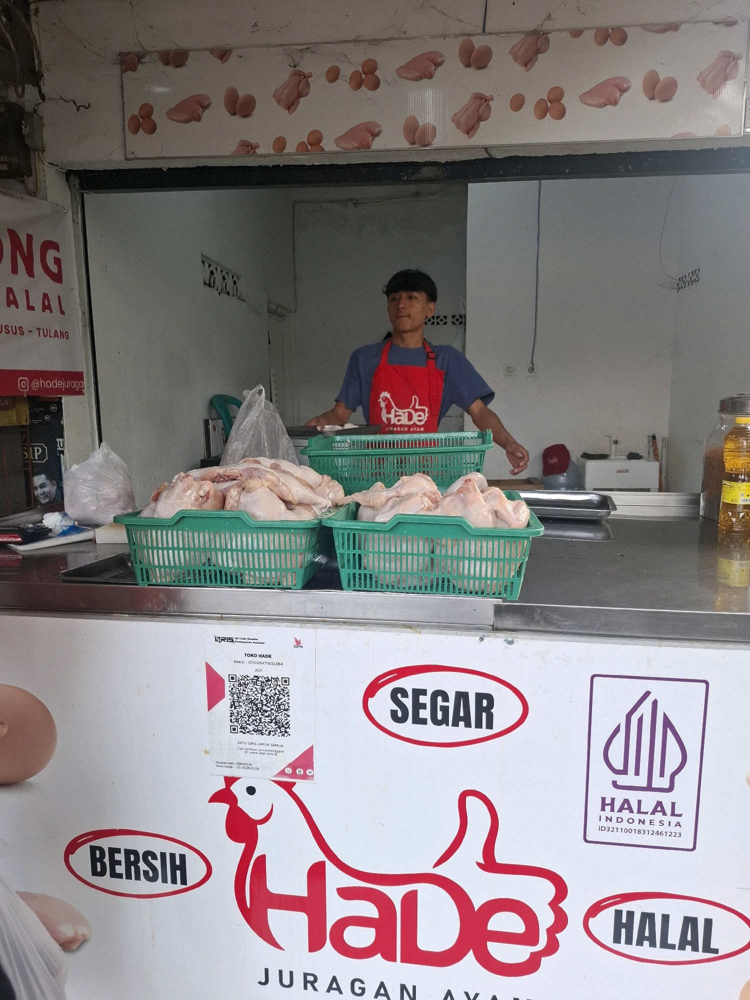A butcher shop in Bojong, Indonesia, displays raw chicken in green baskets on a metal counter. A young male employee wearing a blue shirt and a red apron stands behind the counter, his right hand extended as if serving. To the left, a sign reads "ONG LALAL SUSU - TULANG" with a social media handle "@hadejuragan" below. To the right, a bottle of cooking oil stands next to a container. The shop features a mural with illustrations of chicken parts and eggs. A QR code for "TOKO HADE" is visible on the counter. Text on the counter includes "SEGAR," "BERSIH," and "HALAL" in red and black lettering. The overall scene depicts a typical Indonesian butcher shop environment.