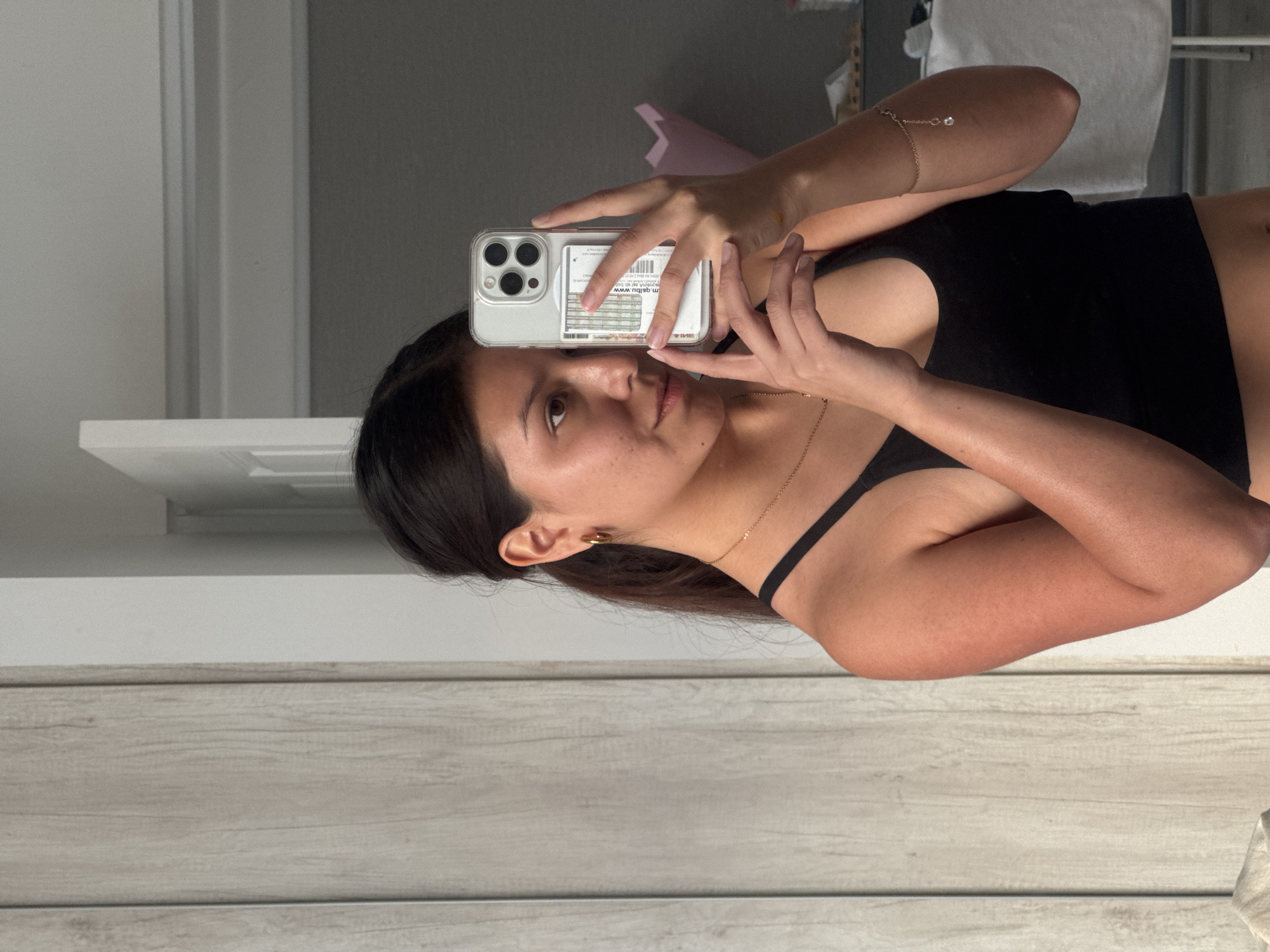 A woman is taking a selfie in front of a light gray wood grain wall. She is wearing a black crop top and has a gold necklace and bracelet. Her phone has a clear case with cards in the back. The location is not evident in the image to confirm the city and country.
