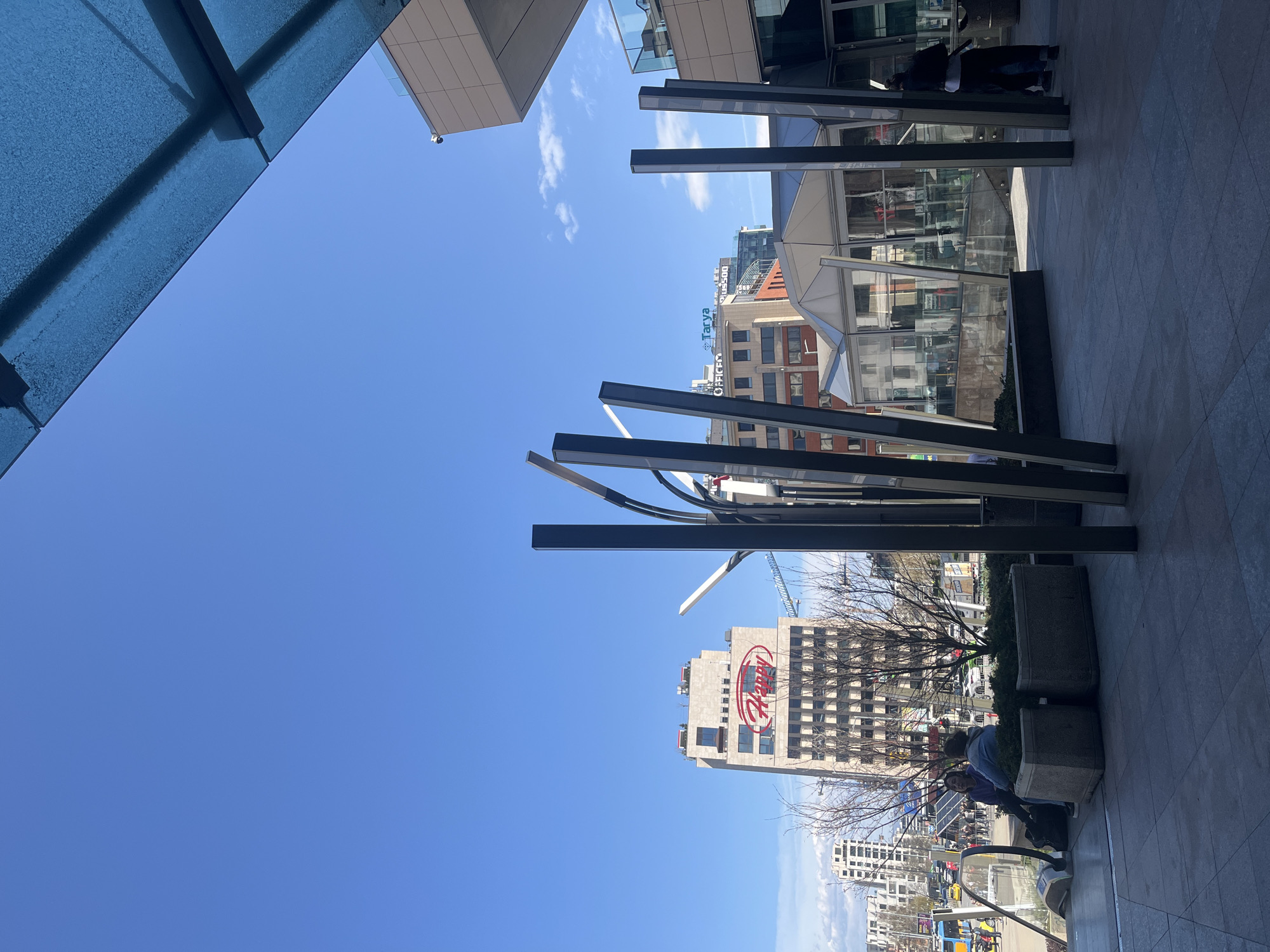 The image shows an urban scene in Sofia, Bulgaria. The viewpoint is from a pedestrian walkway looking towards buildings across the street.
Architecture: The buildings visible include a modern glass and steel structure, and a taller building with a "Happy" logo displayed prominently on its facade. There is also a sign visible on another building reading “Tarya plus500”.
Objects: An artistic metal sculpture is a prominent feature in the foreground, situated between the pedestrian walkway and the street. There are also planter boxes along the walkway.
People: People are walking and interacting near the walkway, but the image does not allow for detailed recognition.
Environment: The weather appears to be sunny and clear, with a bright blue sky. A construction crane can be seen near the "Happy" building.