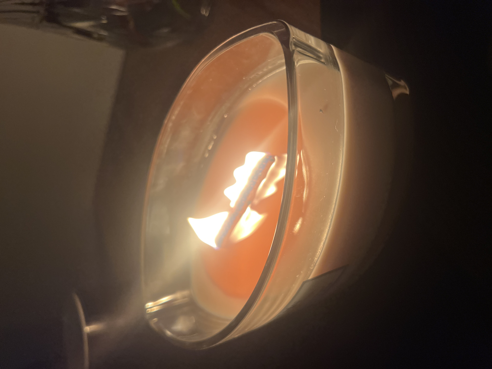 The image shows a close-up of a lit candle in a glass container. The candle wax is a light peach color, and the surface of the wax appears to be slightly melted, with a brighter orange layer visible just beneath the surface around the wick. The wick itself is burning with two distinct flames, casting a warm glow. The glass container has a curved, boat-like shape and is transparent, revealing the wax within. The background is dark and out of focus, suggesting an indoor setting with dim lighting. There are no people or animals visible in the image, nor any specific landmarks that would indicate a location in Vilnius, Lithuania. The overall impression is one of a cozy, tranquil atmosphere.