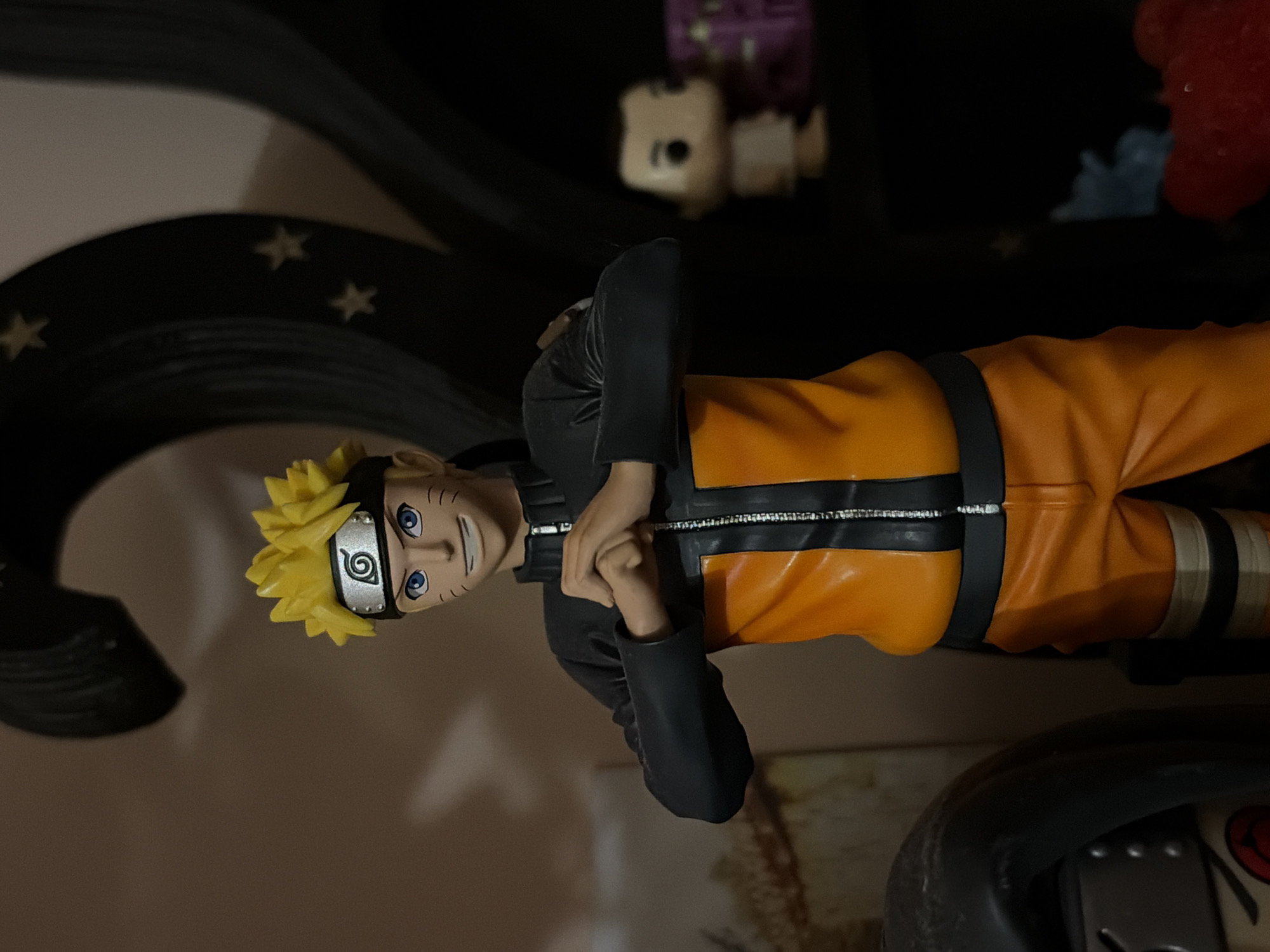 The image features a Naruto figurine, dressed in his signature orange and black outfit, with his hands clasped in front of him. It is positioned on a shelf with other figurines and decorative items visible in the background. The background includes a dark arch-shaped decorative element with star-like patterns. No information suggests the presence of people or that the image was taken in Varna, Bulgaria.