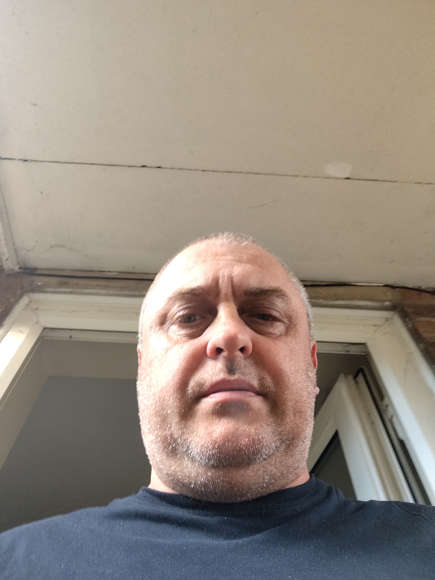 A close-up selfie shows a middle-aged man from a low, upward-looking angle. His head occupies the central portion of the image, and he has fair skin, a shaved or very short haircut, and light-colored stubble on his face. He is wearing a dark, possibly black, crew-neck t-shirt. His expression is neutral.
Above the man's head, a light-colored, likely off-white, ceiling or overhang is visible. Below this, a portion of a robust door or window frame, possibly made of wood or similar material, is seen. The frame features darker sections, suggesting brickwork or detailing, and a thin, dark line, possibly a cable or pipe, runs along its top edge. The area visible through the opening appears dark, indicating an interior or shaded space.
The scene suggests an outdoor or semi-enclosed architectural setting, such as a porch or entryway, within Huntingdonshire, United Kingdom. The lighting indicates it is daytime, with soft illumination that suggests an overcast sky or a shaded position. There is no visible text in the image.