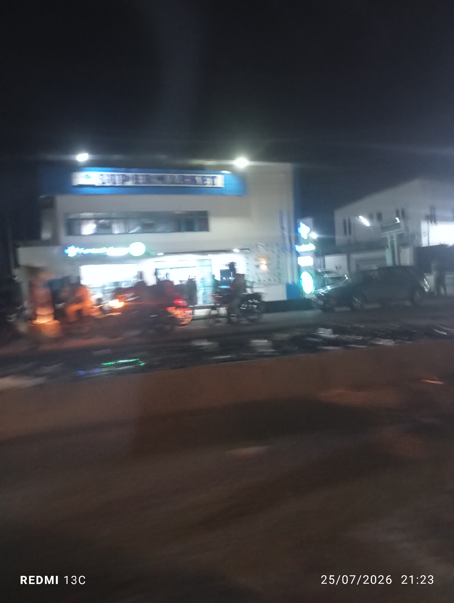 An exterior, night-time, blurred photograph shows a brightly lit supermarket with a blue and white sign that reads "SUPERMARKET" in white lettering against a blue background. To the left of the supermarket entrance, a person is visible, appearing to be standing next to a cart or a bicycle. Further to the left, there is a bright orange glow, possibly from streetlights or a fire. A dark-colored SUV is parked to the right of the supermarket, and several people are standing near it. Another building, lighter in color, is visible to the right of the supermarket, with windows reflecting light. The foreground shows a wet surface, possibly a road or pavement, with reflections of lights. The bottom left corner of the image displays the text "REDMI 13C", and the bottom right corner shows the date and time "25/07/2026 21:23". The scene appears to be in a commercial area of Abeokuta, Nigeria.