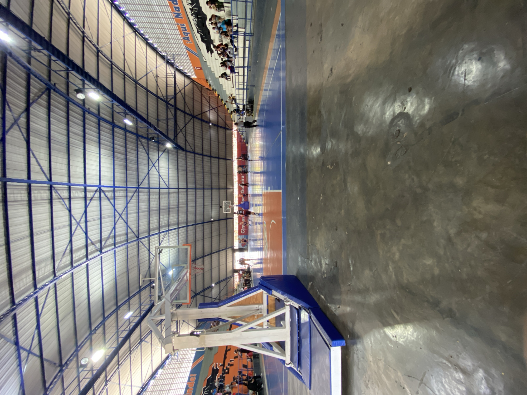 The image shows an indoor basketball court within a gymnasium in Valencia, Venezuela. The court is blue with orange accents. A basketball hoop is visible on the left side of the frame. Spectators are seated in the bleachers in the background, which are decorated with an orange and black mural and text that reads "¡Aquí Nada!". Several individuals, presumably players and possibly a referee, are on the court, engaged in what appears to be a game. The gymnasium has a distinctive arched metal roof with blue beams and is illuminated by overhead lights. The floor in the foreground is a dark, polished concrete.