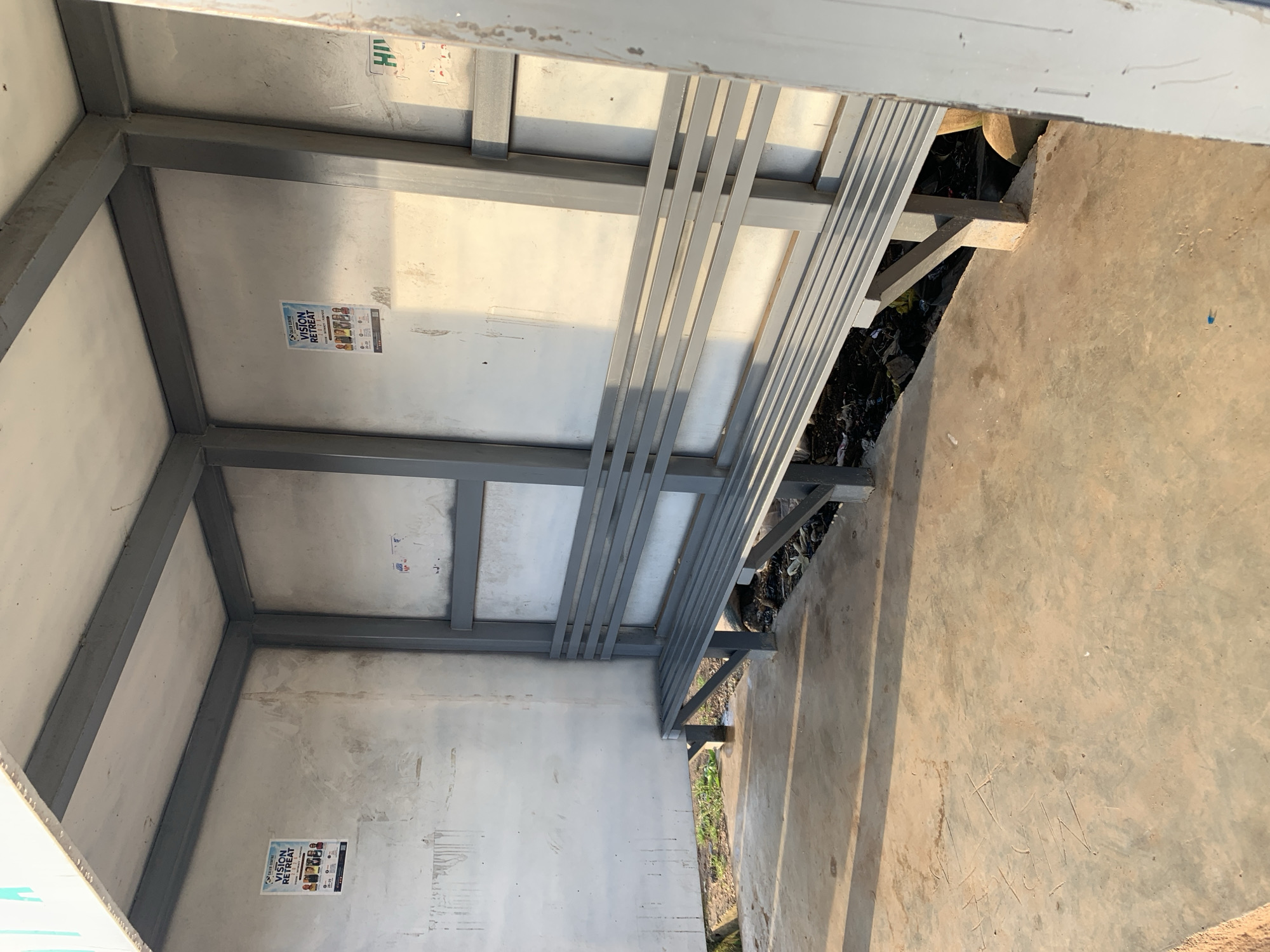 The image depicts an outdoor bus stop shelter in Rafin Par, Nigeria. The shelter is constructed with a grey metal frame and has a corrugated metal roof. The walls appear to be made of rough, unfinished concrete. A long, grey metal bench with several slats runs along the back of the shelter. The ground inside and outside the shelter is concrete. Sunlight casts shadows on the ground, indicating it is likely daytime. There are two posters visible on the left wall, advertising a "VISION RETREAT" with dates and images of people. There are also some handwritten markings on the concrete floor. In the lower right portion of the image, a section of the shelter's right wall is visible, also made of metal. The area around the bus stop appears to be unkempt, with some debris visible on the ground beneath the bench.