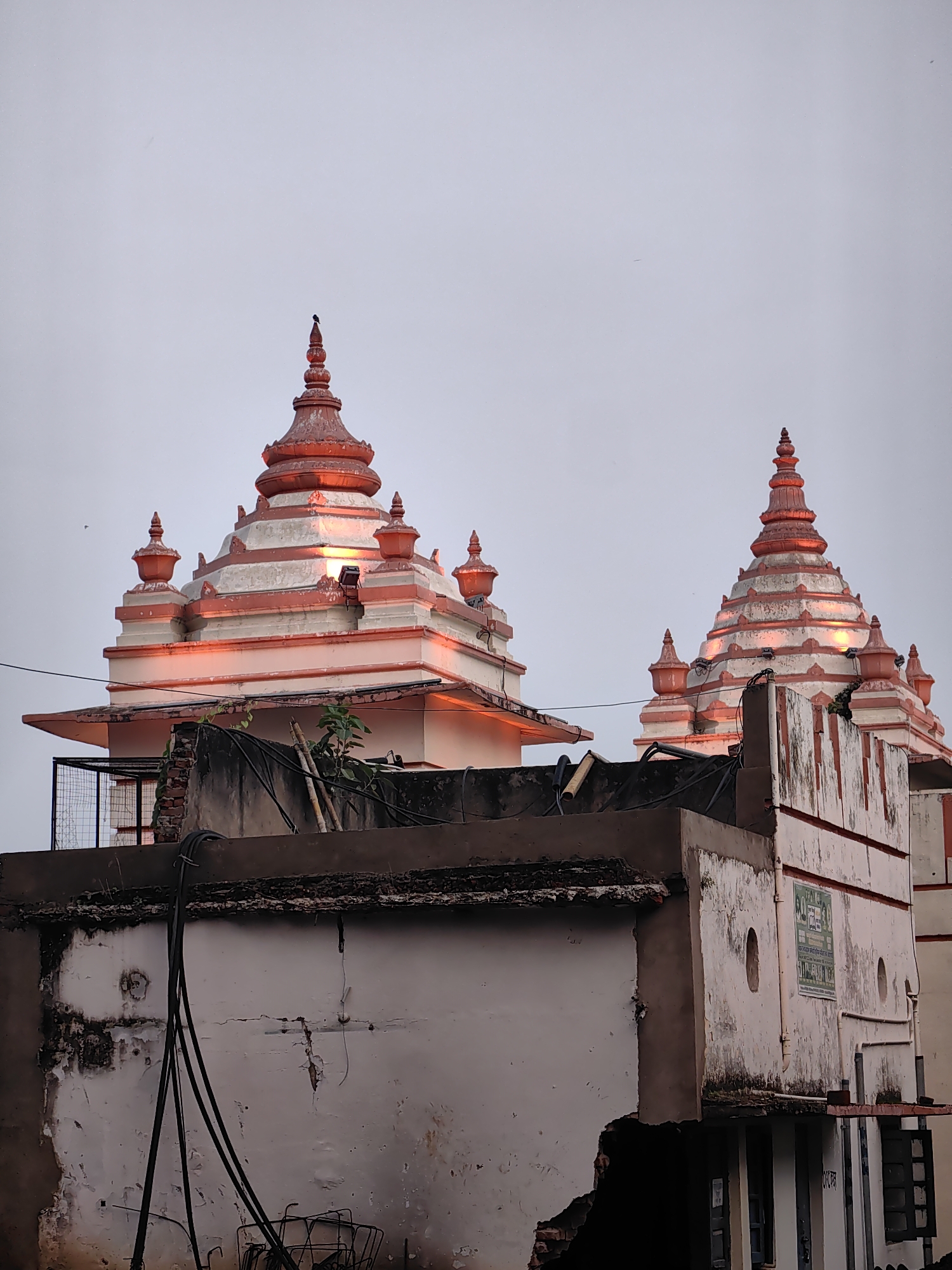 A medium shot captures the upper portions of two intricately designed temple spires, illuminated by warm orange light, against a pale, overcast sky. The spires, adorned with stacked levels and smaller finials, are predominantly white with red accents. Below the temples, the edge of a weathered, dilapidated building is visible, featuring cracked plaster, exposed brickwork, and a network of black electrical wires strung across its surface. The building's facade is marked by peeling paint and grime, suggesting neglect. A metal security fence can be seen on the left side of the image, partially obscuring the temple's lower levels. In the far distance, a tiny, dark bird is perched atop the left spire. The scene is static, with no discernible human activity or interaction. The overall impression is one of aged architecture juxtaposed with the encroaching decay of the foreground structures, all under a muted sky. The image was captured in Varanasi, India, a city known for its numerous temples and ancient religious sites.