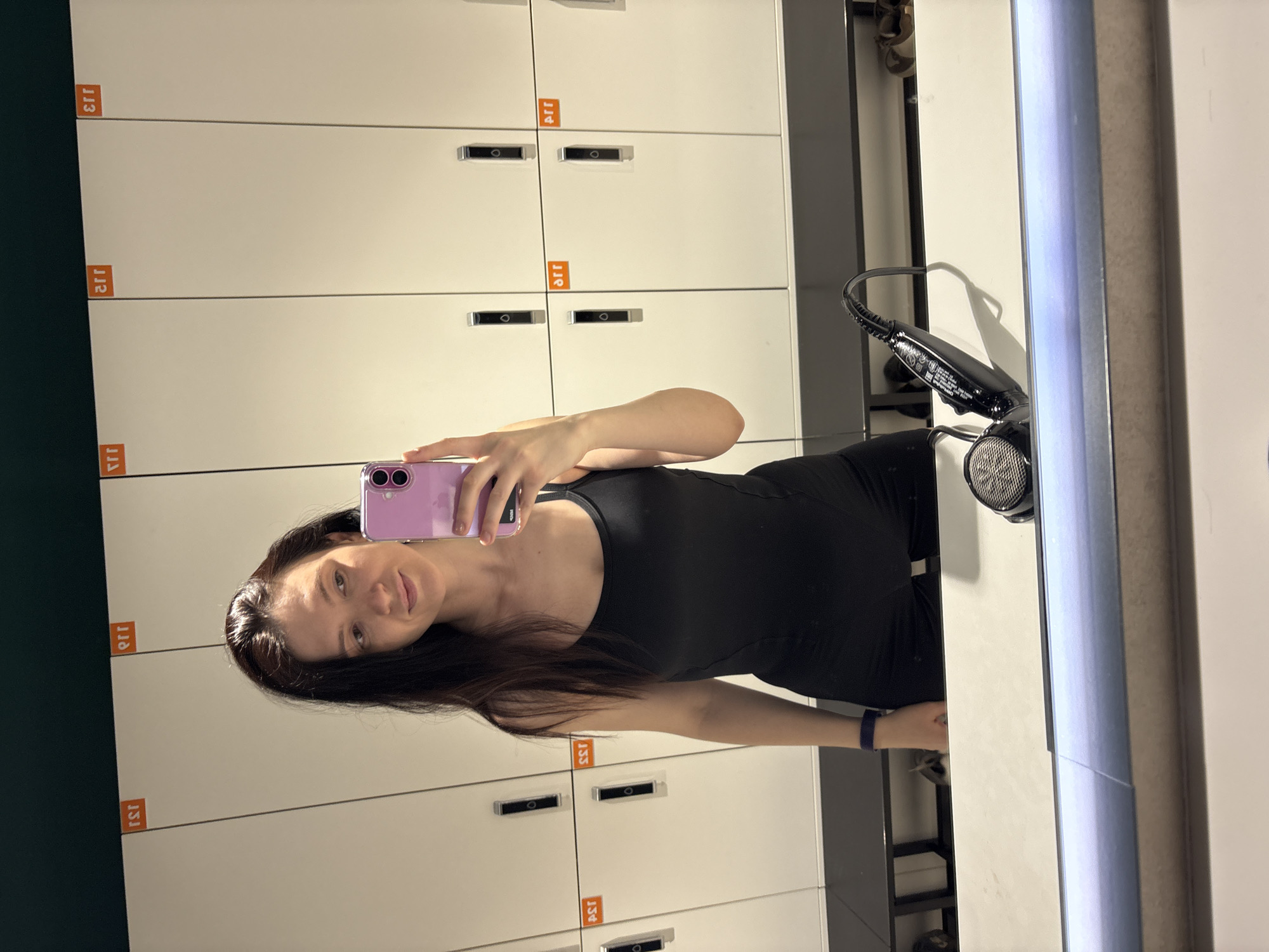 A young woman is captured taking a mirror selfie in a well-lit locker room, likely in Kazan, Russia. She has long dark hair and is wearing a black sleeveless top or dress, with a dark blue or purple wristband visible on her left wrist. She holds a light purple smartphone, equipped with a distinct camera module, to capture her reflection. Her expression is calm, featuring a gentle smile.
The background of the image, visible through the mirror, consists of several tiers of white lockers. Each locker features a dark, circular handle and an orange rectangular label with numbers. Due to the reflection, these numbers appear reversed, reading as '113', '112', '111', '114', '115', '133', and '134'. To the right of the woman, also reflected in the mirror, a black hairdryer rests on a counter or shelf. The indoor lighting is bright and even, indicating a clean and functional environment typical of a gym or sports facility. The scene depicts a casual self-portrait moment.