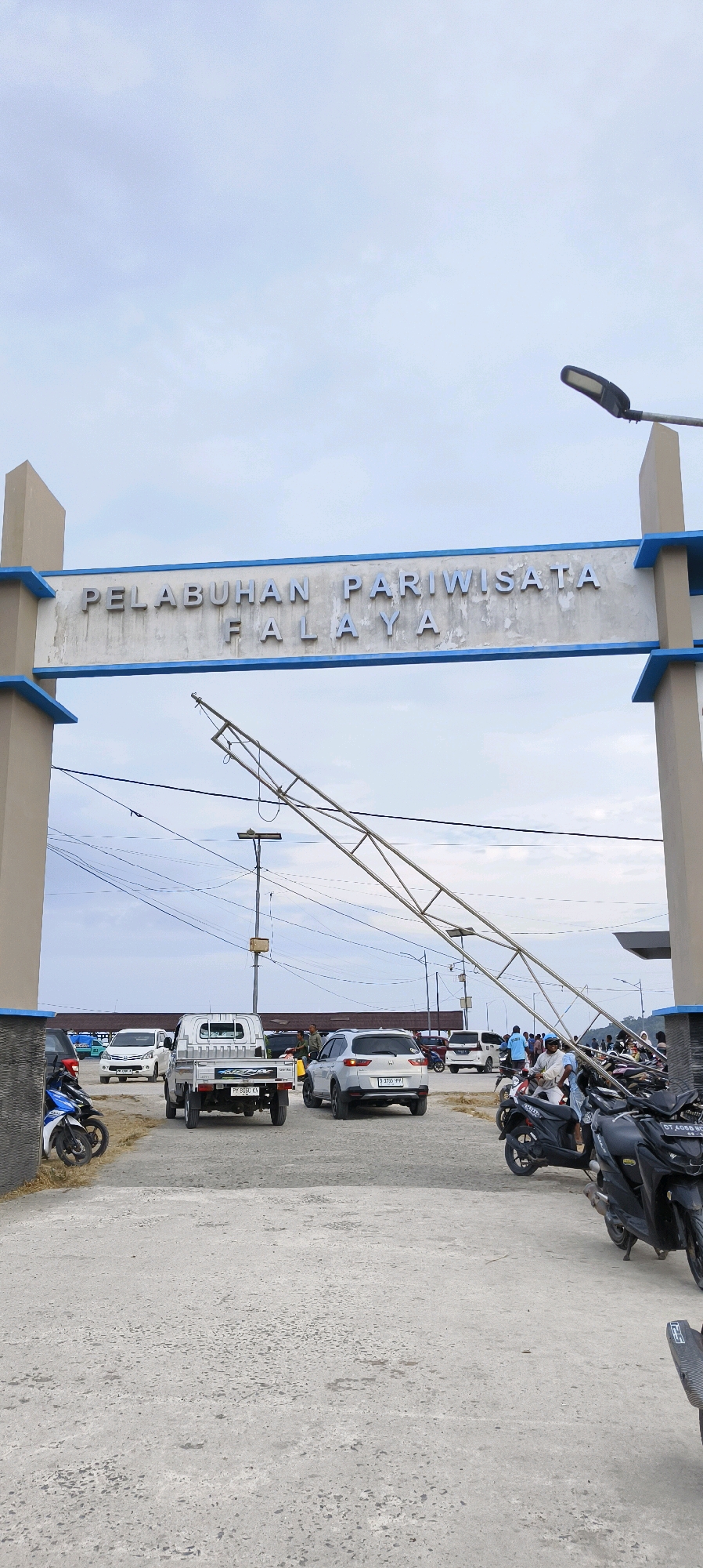 This is a photograph taken outdoors in Waisai, Indonesia, featuring an entrance archway labeled "PELABUHAN PARIWISATA FALAYA." The arch is supported by two substantial pillars on either side, with the text mounted on a horizontal beam between them. A metal structure, possibly part of a larger construction or a damaged element, is angled across the frame from the upper left to the lower right. The ground is paved with concrete.
Several vehicles are present, including a white pickup truck in the center foreground, a white SUV to its right, and various motorcycles parked and on the road. Some individuals are visible, including people near the vehicles and in the background. The sky is overcast with a pale blue hue. The scene appears to be a port or tourist area, indicated by the signage and the presence of transportation.