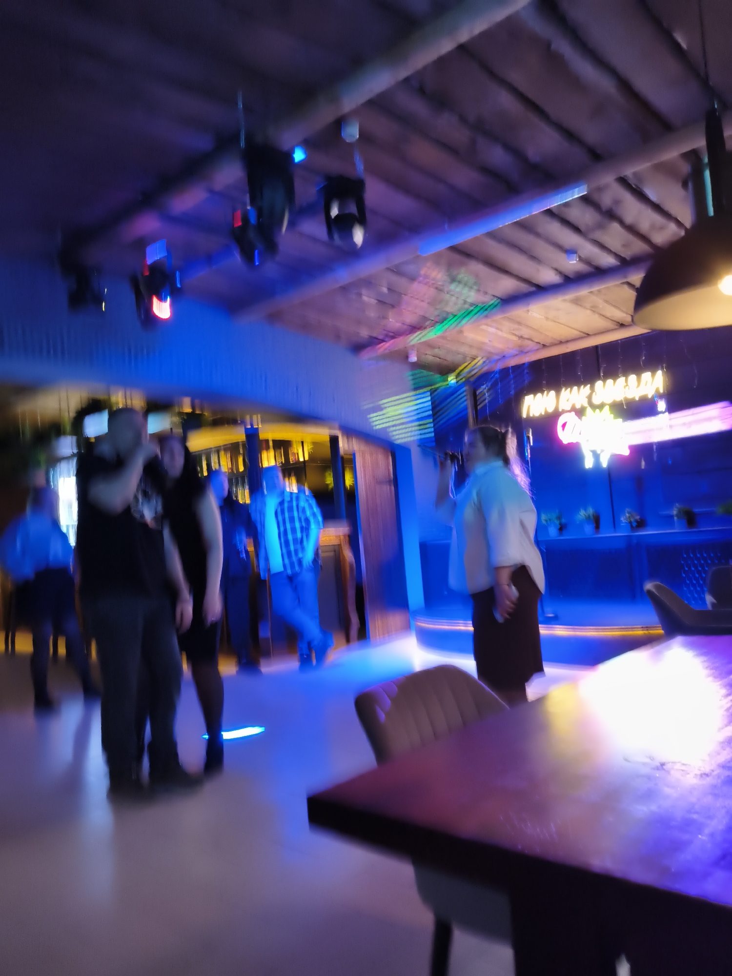 The image, captured indoors and appearing somewhat blurry due to low light or motion, depicts a lively scene inside a nightclub or karaoke bar in Chaikovsky, Russia. The setting is characterized by a distinctive wooden slat ceiling and a vibrant, albeit dim, lighting scheme dominated by blue hues from LED strips and spotlights.
Several individuals are visible throughout the space. In the mid-ground on the right, a person wearing a light-colored shirt and a dark skirt is singing into a microphone on what appears to be a small stage area. Behind them, a prominent neon sign glows with Cyrillic text, clearly reading "ПОЮ КАК ЗВЕЗДА" (I sing like a star) in white, accompanied by another less clear pink/purple phrase beneath it.
To the left, a group of people stands, seemingly socializing or watching the performance. One man in a black t-shirt and dark trousers, and another in a plaid shirt, are discernible, though their faces and details are indistinct due to the blur. Further in the background, another man in a light blue shirt is visible near what appears to be a bar or service counter.
The floor is light-colored, reflecting some of the intense blue lighting. Spotlights emitting red, blue, and green light are mounted on the ceiling beams, casting dynamic patterns across the room. A dark wooden table and an upholstered chair are partially visible in the bottom right foreground, suggesting seating for patrons. The overall atmosphere conveyed is one of nighttime entertainment and social gathering.