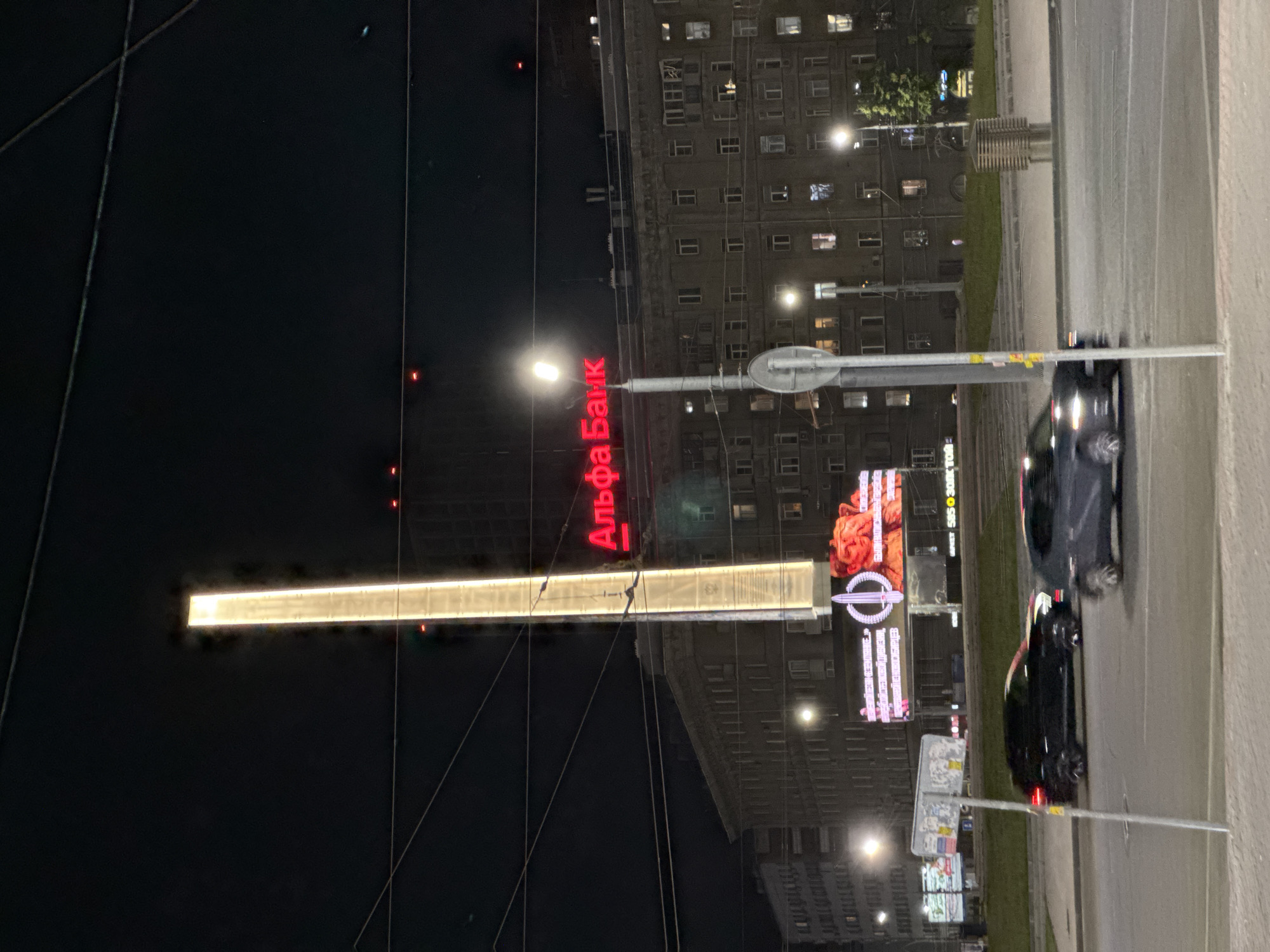 The image shows a night scene in Novosibirsk, Russia, with a tall, illuminated monument or structure dominating the center of the frame. Red neon lettering on a building behind the monument spells out "Альфа Банк," indicating a branch of Alfa-Bank. There are several cars on a street in the foreground, with one black car to the left and a gray car to the right, both in motion. The buildings in the background are lit with windows, suggesting an urban environment. Overhead wires and streetlights are also visible.