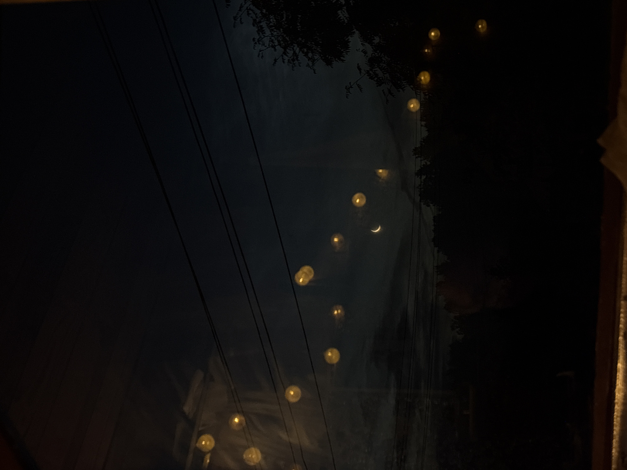 A dark, grainy image captures a nighttime scene, possibly from a window. In the foreground, a blurred wooden surface runs horizontally across the bottom. The background depicts a deep blue-black sky with several horizontal lines that could be power lines. Scattered across the sky are blurry, glowing yellow orbs, likely reflections or lights. Towards the middle of the frame, a faint crescent moon is visible. Silhouetted against the sky is a dark line of trees and foliage. To the right, the dark silhouette of a tree with visible branches stands out. The overall impression is one of a dimly lit, possibly rural or suburban, outdoor environment at night. There is no specific indication of the city of Jāņupe, Latvia.