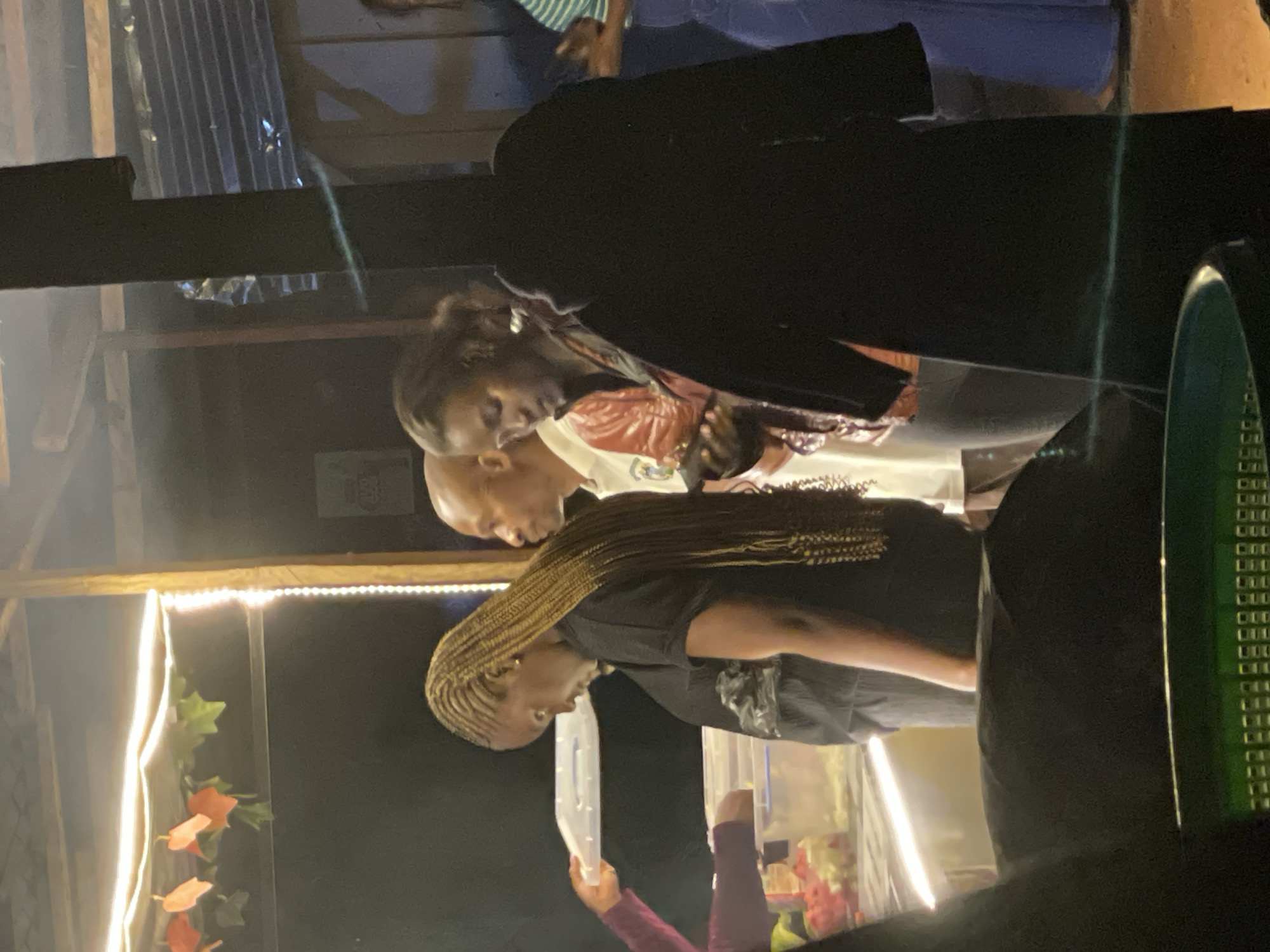 The image shows a dimly lit outdoor gathering. In the foreground, a person wearing a black jacket is largely obscured by shadow. Behind them, a young woman with braided blonde hair is looking down at her phone. To her left, a bald man in a white shirt is visible. Further left, a person's arm is holding up a white plastic container. In the background, there is a thatched roof structure illuminated by string lights. The scene appears to be set in Durumi, Nigeria, possibly at a community event or informal celebration.