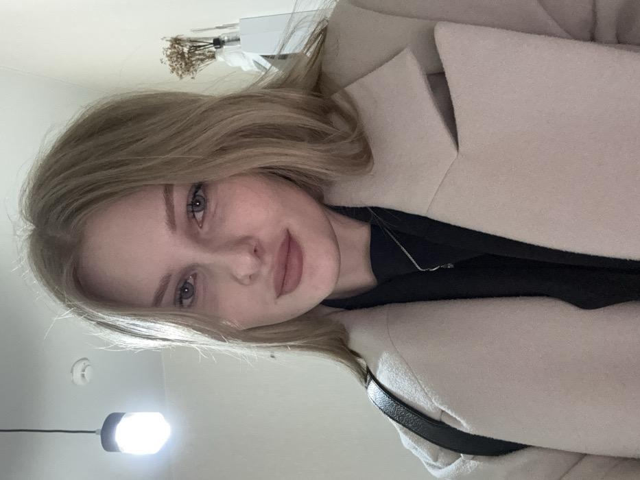 A female subject, appearing to be a young adult with light skin, blonde hair, and blue eyes, is depicted in a close-up, head-and-shoulders shot. She is wearing a light beige coat with lapels over a black garment, accessorized with a thin silver necklace featuring a small, dark pendant. A black, shiny strap, consistent with a bag, is visible over her left shoulder. Her hair is parted and frames her face. Her lips are a light pink/nude shade, and she maintains direct eye contact with the camera, displaying a neutral to slightly smiling expression. The background consists of plain, light-colored walls. To the upper left, a luminous white cylindrical light fixture hangs from a black cord. To the upper right, a white shelf or surface is partially visible, holding a clear vessel containing several dried plant stems. The specific location within Perm, Russia, cannot be determined from the visual information presented in the image.