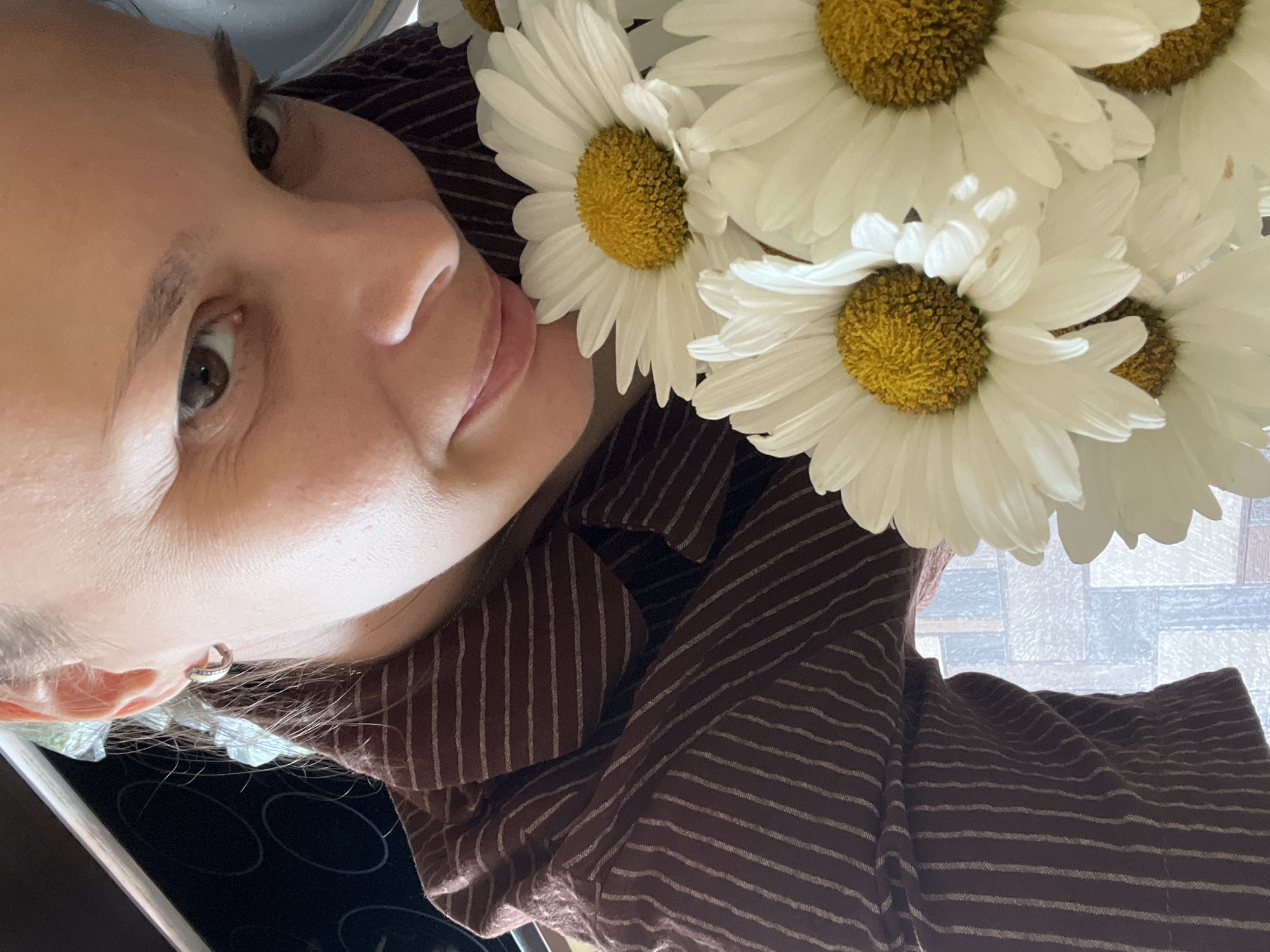 A close-up selfie shows a woman holding a bouquet of white daisies with yellow centers. She is wearing a dark brown shirt with thin, light-colored vertical stripes and a silver hoop earring in her visible left ear. The woman has brown eyes and is looking directly at the camera with a slight smile. The background is blurred, showing hints of a kitchen appliance on the left and a window with a patterned glass on the right. The setting appears to be indoors. The image does not contain any information that would indicate a specific location in the city of Tolyatti, Russia.