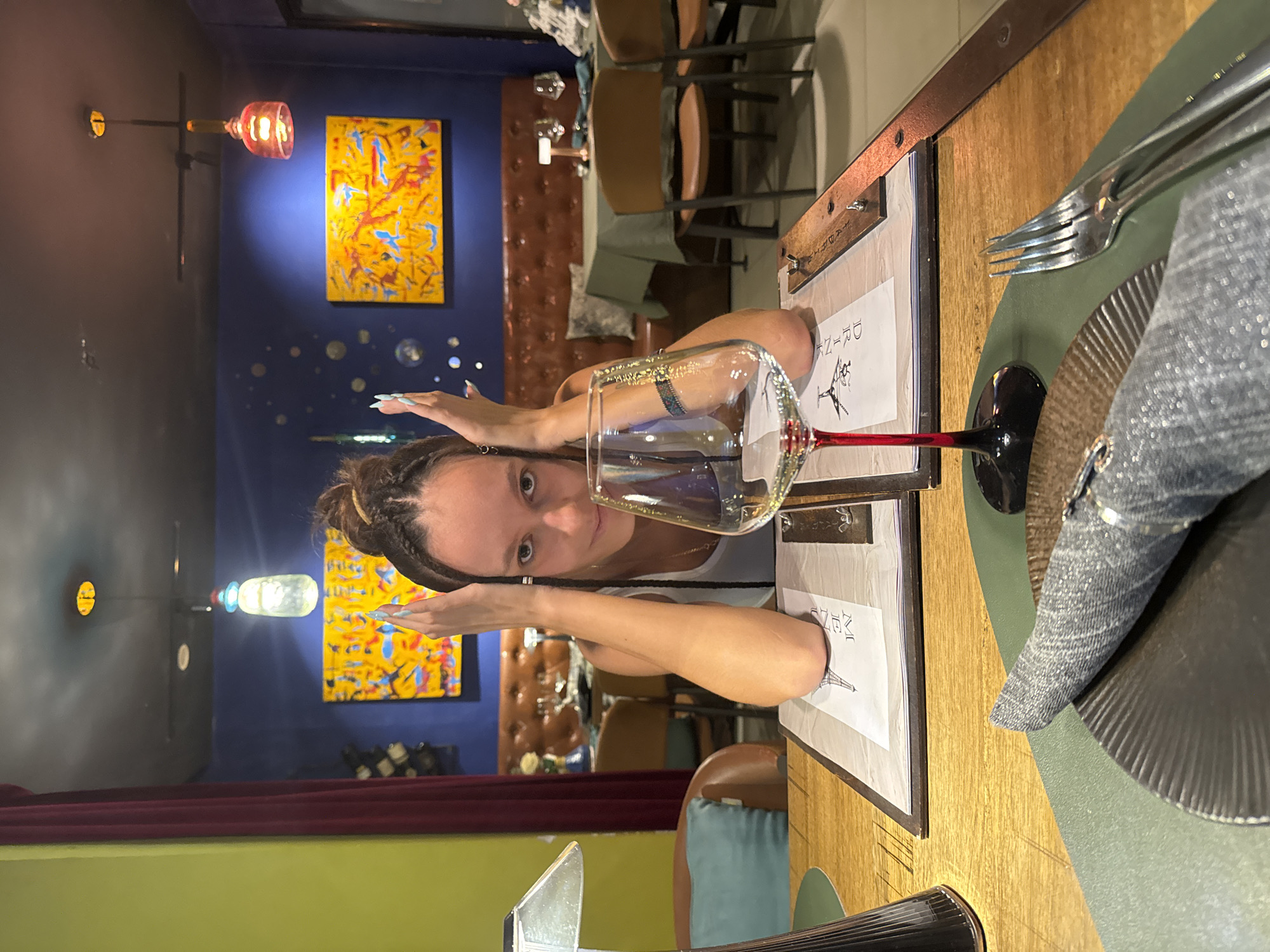A woman is sitting at a table in a restaurant, resting her forearms on a menu. She is looking at the camera with her hands on either side of her head. In front of her is a wine glass, a plate, a fork, and a napkin. The restaurant has blue walls with yellow paintings and brown leather booths. The image was taken in Hanoi, Vietnam.