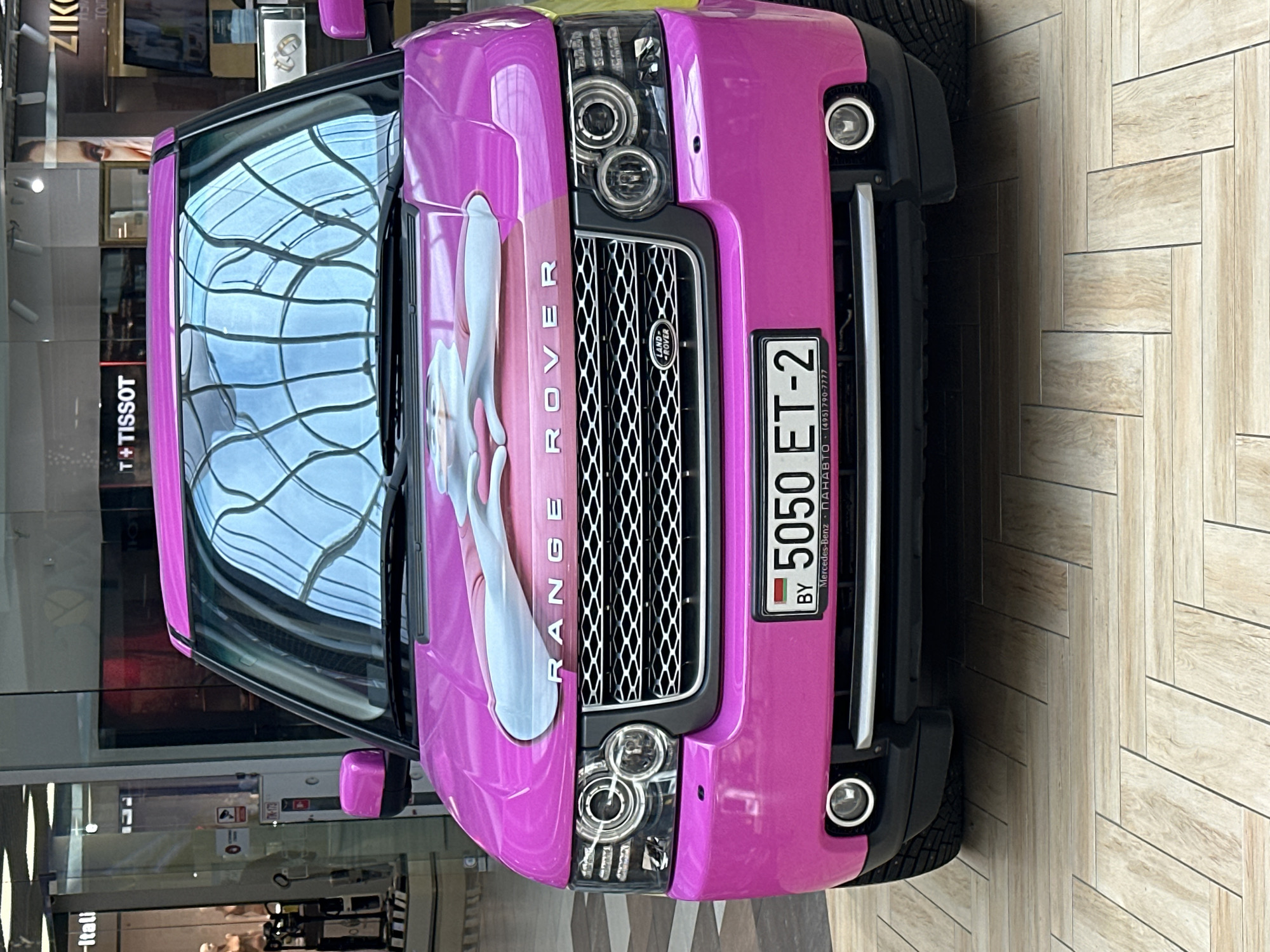 A bright pink Range Rover is parked indoors on a tiled floor. The car features a cartoonish graphic on its hood. The license plate reads "5050 ET-2 BY". In the background, a storefront with "T+ TISSOT" signage is visible, along with reflections of a glass ceiling. The scene appears to be in a shopping mall.
