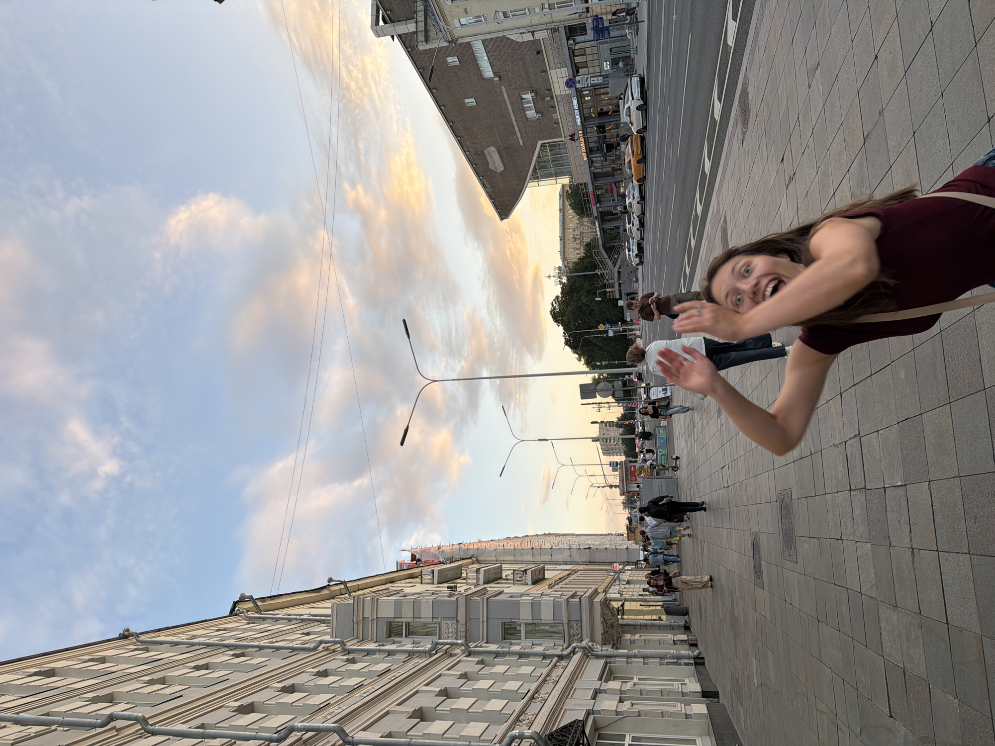 A young woman with long brown hair, wearing a maroon top and a beige strap across her chest, is captured in a full-body, low-angle shot with an exaggerated, excited facial expression and her arms raised in a welcoming gesture. She is standing on a stone-tiled sidewalk in front of a street with parked cars and buildings. The scene appears to be in Moscow, Russia, given the architectural style of the buildings, particularly the ornate facade of the building on the left, which is typical of pre-revolutionary or early Soviet architecture. The sky is a pale blue with wispy, golden-pink clouds, indicating either sunrise or sunset. The street is lined with lampposts and various shops. In the background, there is a large, angular, modern building with signs for "BURGER HEROES" and "HOT DOG BLDG." The overall ambiance suggests a lively urban environment during golden hour.