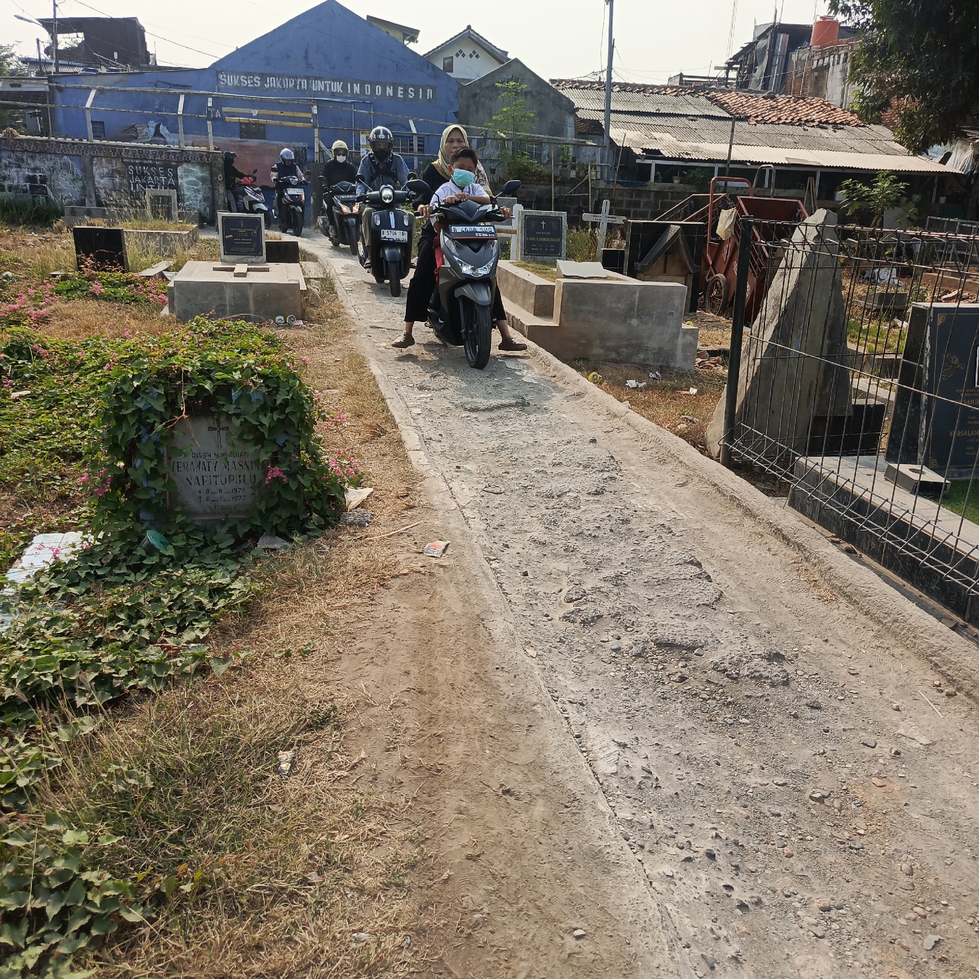 The image depicts a cemetery in South Jakarta, Indonesia, under a clear sky. A dirt path runs through the cemetery, lined with graves and overgrown vegetation. Several people are seen riding on motorcycles, navigating the uneven terrain. Some individuals are wearing masks, and one person appears to be a child. In the background, buildings with blue and corrugated metal roofs are visible, along with a fence and some construction materials. The overall scene suggests a solemn yet ordinary day in this urban cemetery.