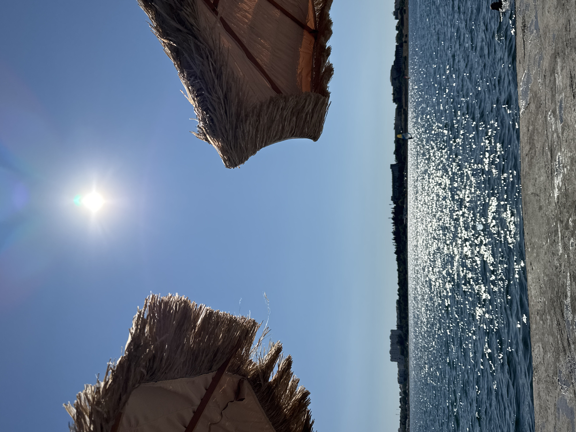 The image depicts a sunny day at the Sevastopol seaside. Two thatched umbrellas frame the shot from the left and right, casting shadows on the viewer. The bright sun is visible in the upper center of the frame, its rays creating a glittering effect on the calm blue water. In the distance, a coastline with buildings and trees stretches across the horizon. There is a hint of a person in the water on the right side of the frame. The foreground is a concrete surface, possibly a pier or promenade, with patches of water. The scene conveys a peaceful and summery atmosphere.