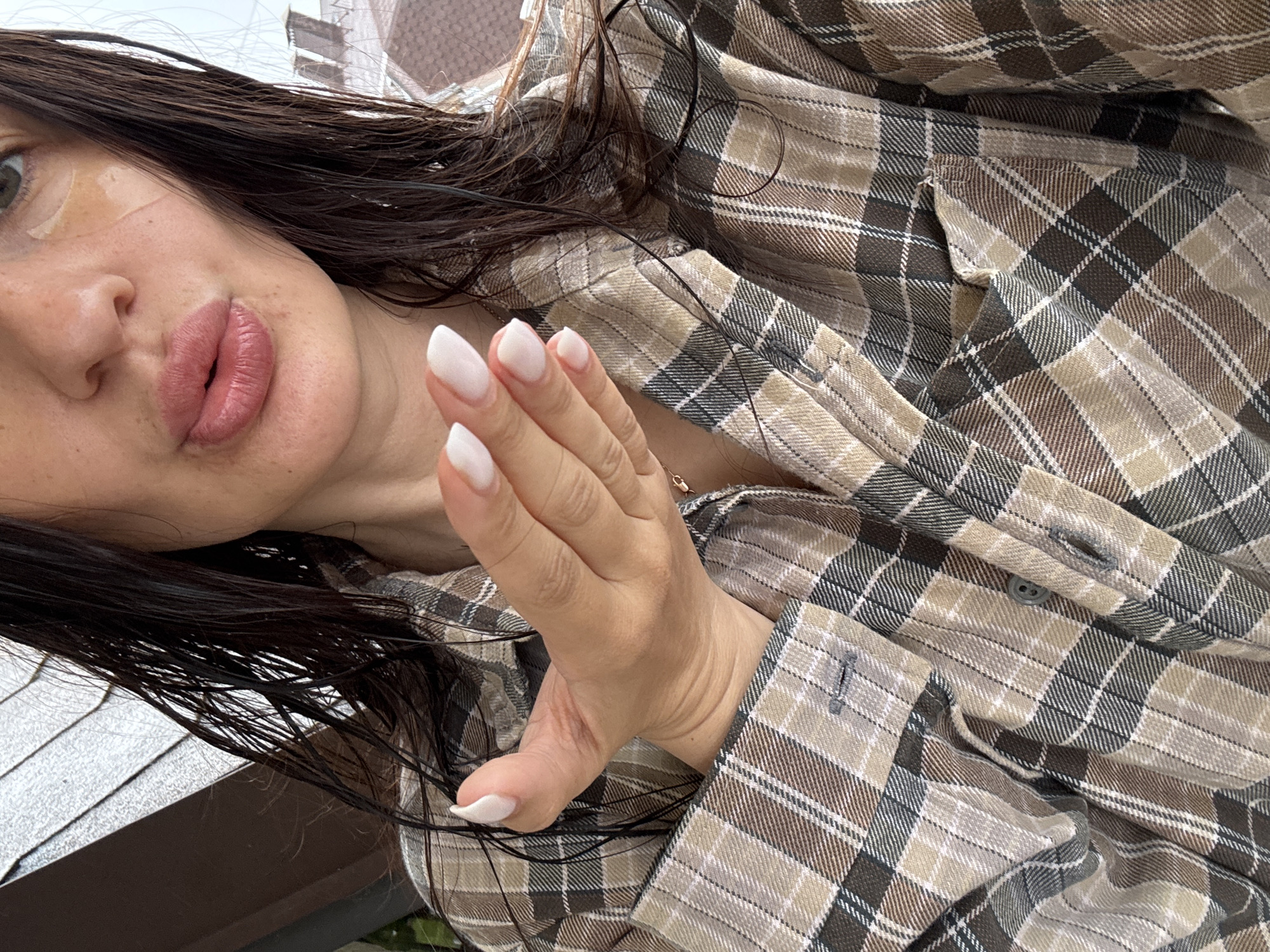 A medium-shot, close-up photograph features a person wearing a brown and beige plaid shirt, positioned at an oblique angle. The person's face, predominantly featuring lips and a portion of the cheek, is visible. There's a small, transparent adhesive patch on the cheek. The hair appears dark and wet. The person's hand, adorned with long, white almond-shaped nails, is raised with the fingers extended.
In the background, indistinct architectural elements suggest a cityscape. Based on the style of architecture, the photograph may have been taken in Sochi, Russia, but additional visual data or metadata would be needed to confirm this.