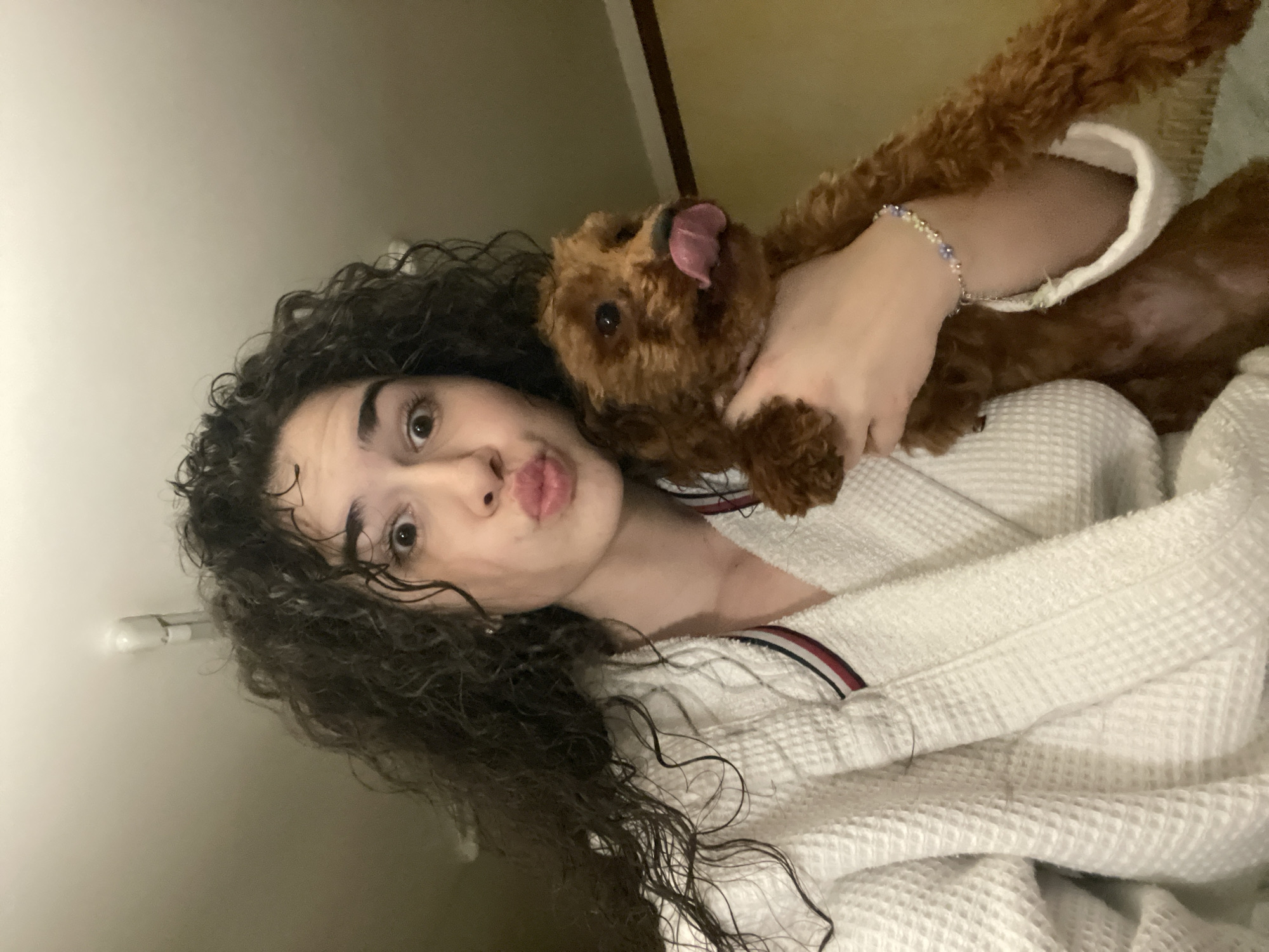 A young woman with curly dark hair, wearing a white waffle-patterned bathrobe, is holding a small, brown poodle. The dog is licking its nose. The woman is making a kissing face towards the camera. The location is unspecified; there is no information to indicate it is in Sofia, Bulgaria.