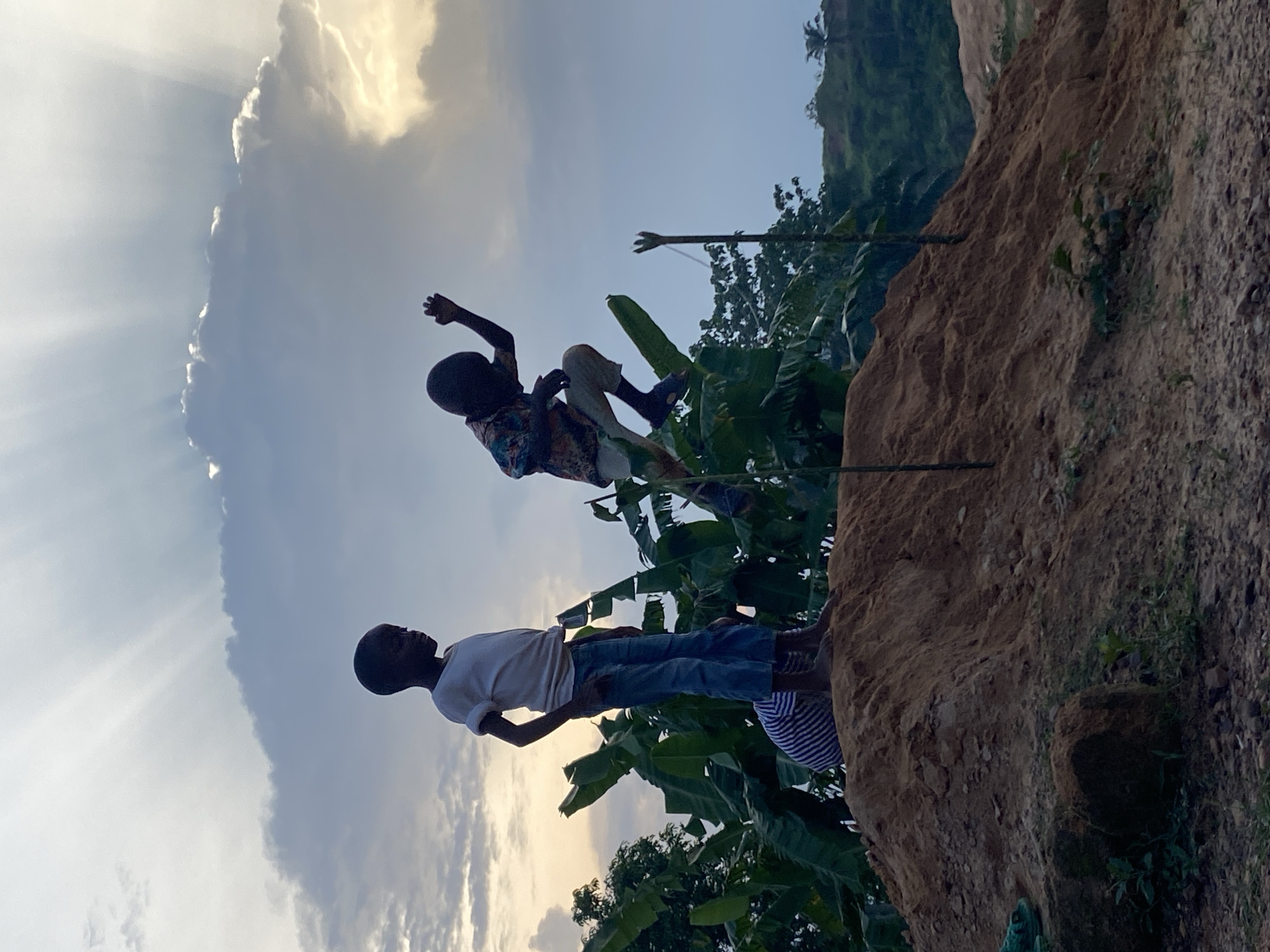 Two children are playing atop a dirt mound in Ogbu, Nigeria. The child in the foreground wears a white t-shirt and jeans, standing near the edge of the mound. The second child, dressed in a patterned shirt and pants, is leaping from the mound. Green foliage, including banana trees, is visible on and around the mound. In the background, there is a cloudy sky with sun rays. The mound appears to be part of a larger dirt structure or hillside.