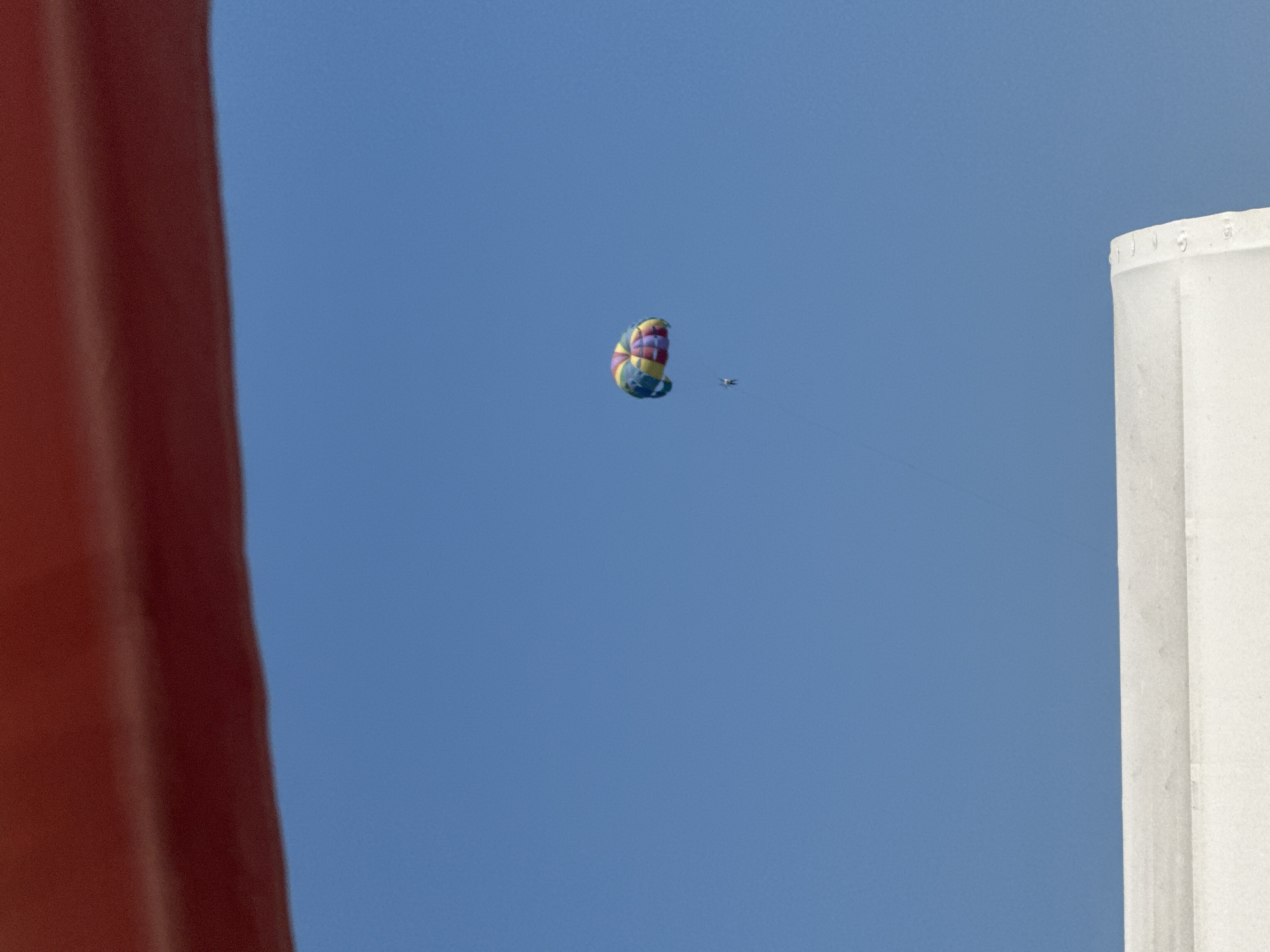 A person is parasailing in the clear blue sky over Sary-Oy, Kyrgyzstan. The parasail is colorful, with red, yellow, green, blue, and purple sections. The person is suspended below the parasail by a rope. In the foreground, the edge of a red structure is visible at the top of the image, and a white structure is visible at the bottom of the image. The white structure appears to be a boat or a dock.