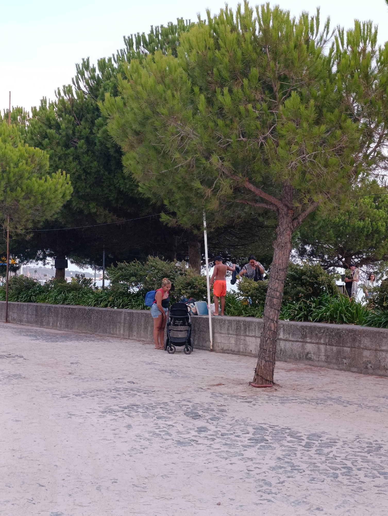 The image depicts a seaside promenade in Setúbal, Portugal, likely during daylight hours. A woman with blonde hair, wearing denim shorts and a dark top, stands beside a black stroller on a cobblestone path. She has a blue backpack slung over her shoulder. In the background, behind a low concrete wall, there are dense green trees and bushes. Further back, several people are visible, including a young boy in orange shorts and two adults near some laundry hanging on a line. The overall scene suggests a relaxed, possibly holiday atmosphere.