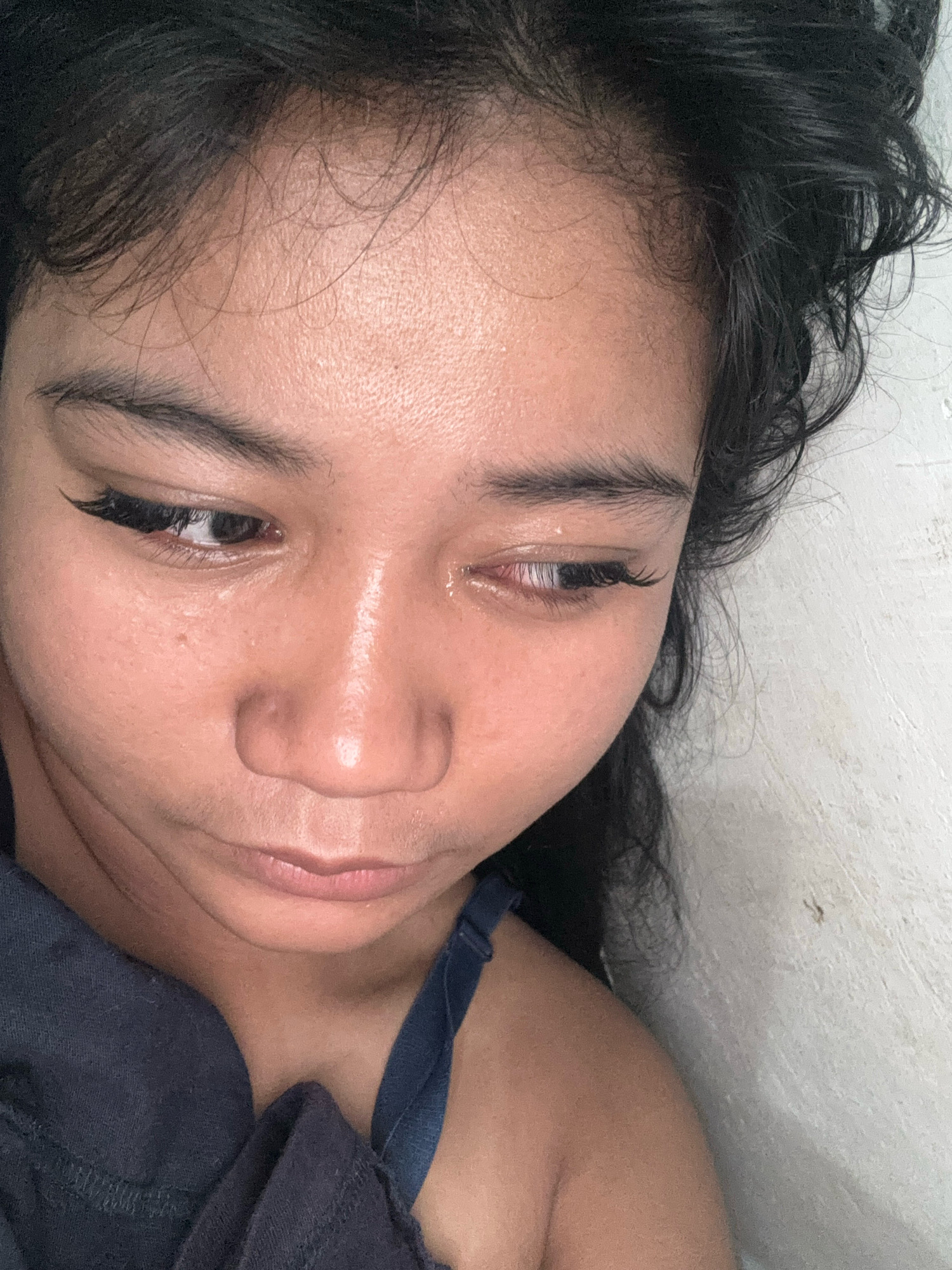 A close-up, eye-level shot shows a young woman's face, from her forehead to her collarbones. Her dark hair is messy and falls around her face. She wears a dark blue bra, visible at the top of her chest. Her skin appears shiny, and her eyes are downcast, with one eye showing a tear. The background is a plain, textured white wall. The lighting is direct and somewhat harsh. The image was captured in North Jakarta, Indonesia.