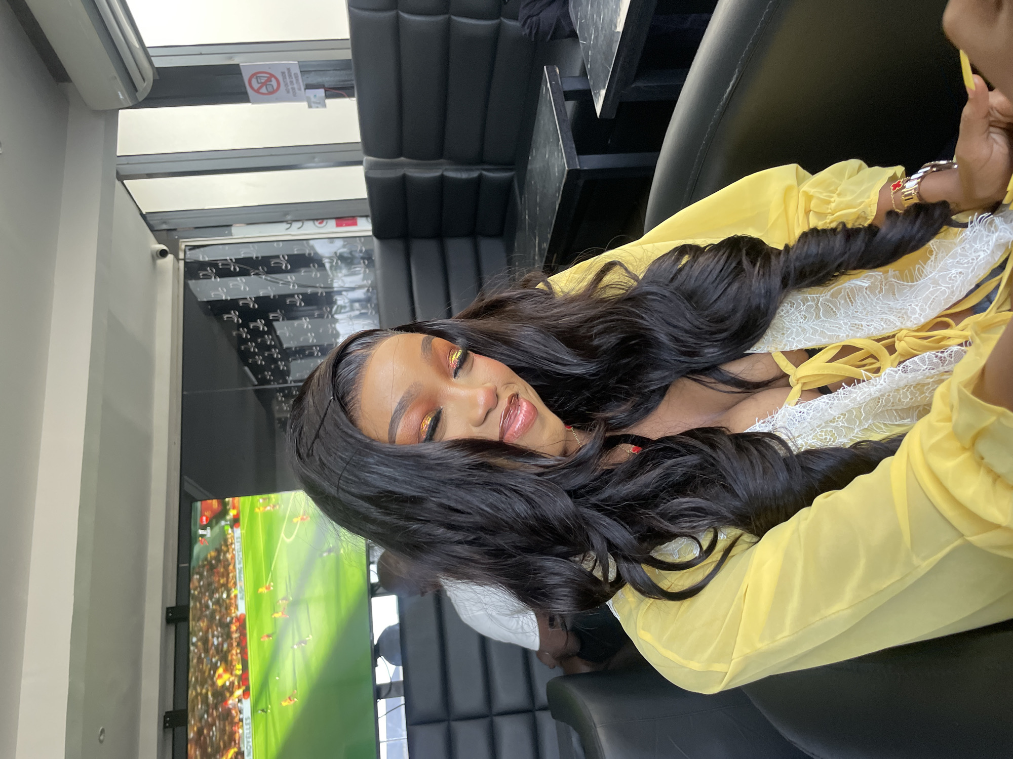 In Animashawun, Nigeria, a woman with long, dark, wavy hair sits in a black leather booth. She is wearing a yellow sheer top with white lace accents. Her makeup includes bold eyeshadow and lipstick. She appears to be looking down with her eyes closed or partially closed. Behind her, a large screen displays a soccer match. The environment suggests an indoor seating area, possibly a bar or lounge, with other patrons visible in the background. The overall impression is a casual social setting.