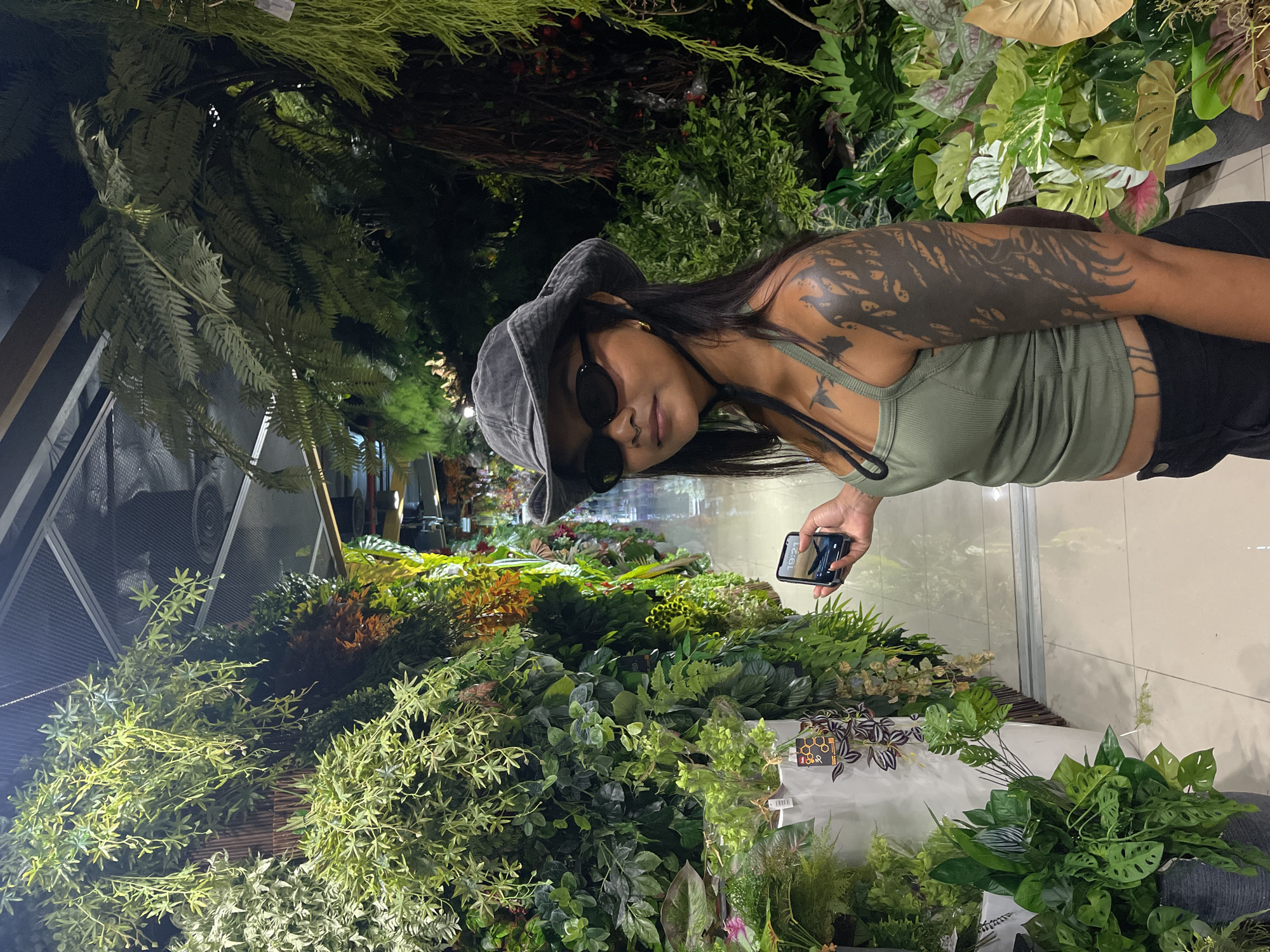 A woman with dark hair, wearing a grey bucket hat, black sunglasses, and a green tank top, stands amidst a dense display of artificial plants. She has extensive black tribal tattoos covering her left arm and shoulder. She is holding a smartphone in her right hand, angled as if taking a picture or recording.
The scene appears to be an indoor plant store or market, filled with a variety of artificial greenery, including ferns, leafy plants, and other foliage. The lighting is dim, suggesting it could be evening or a low-light indoor environment. The background shows more rows of plants and a tiled wall, with a hint of what might be a reflective surface.
The woman is looking towards the camera with a neutral expression. The overall impression is a casual moment captured while browsing or shopping for plants. There is no visible text in the main image, but a small tag with a honeycomb pattern and some Thai script is partially visible on a plant in the lower right quadrant of the image. The orientation of the image is landscape, with the woman positioned in the upper half.