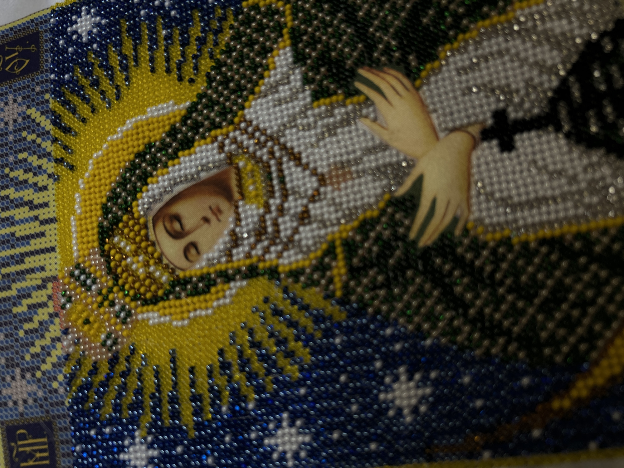An image depicting a representation of the Virgin Mary, possibly a tapestry or beadwork, is shown from an angled perspective. The Virgin's face is visible, surrounded by a halo of yellow and white beads. Her hands are positioned in a gesture of prayer. The background includes blue beads, possibly representing the sky, with white beadwork resembling stars.
The location cannot be determined from the image alone and is not discernible. The city of Cape Coral, United States of America, is not visually referenced or implied in this image.
