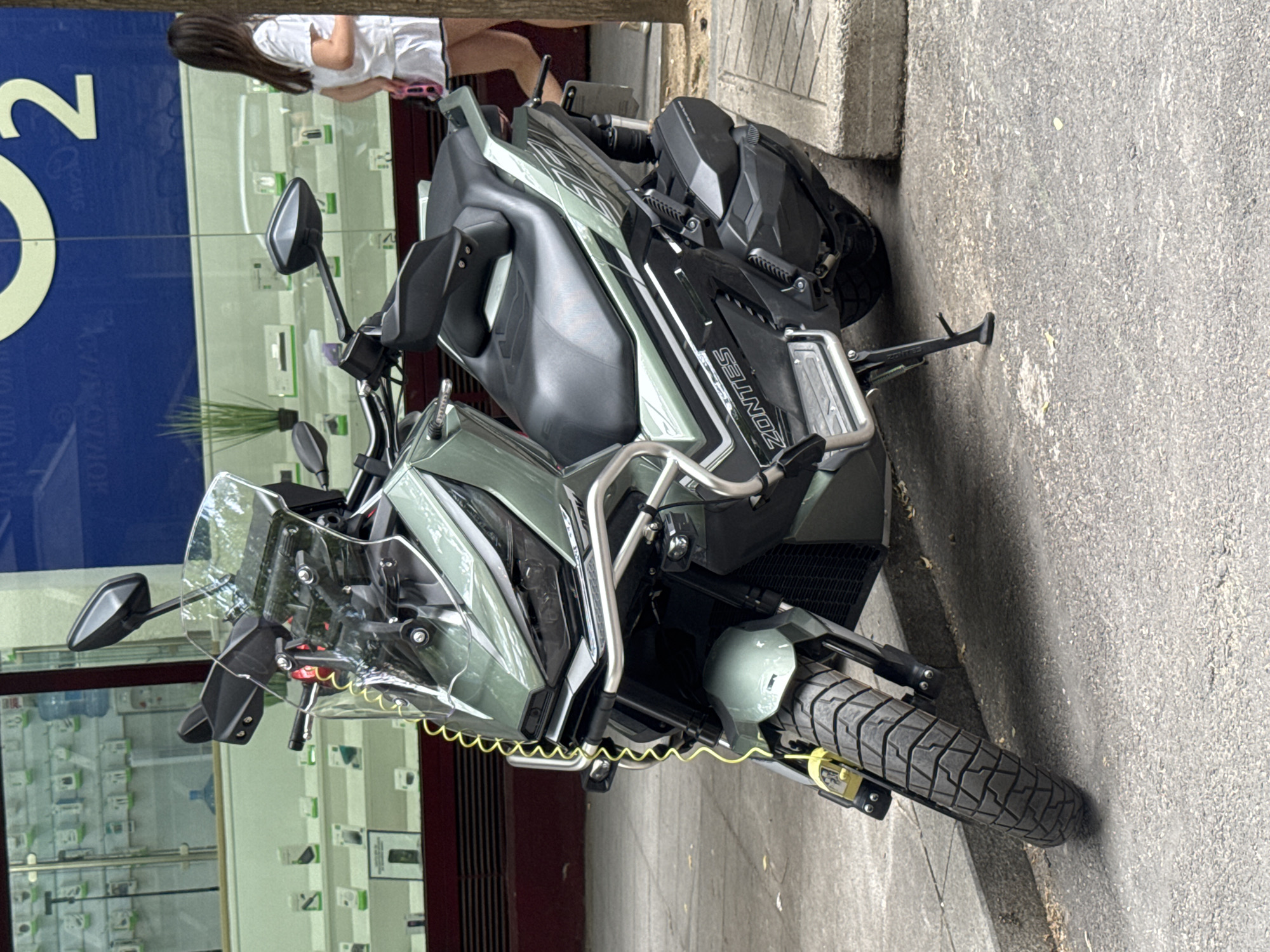 A parked, sage green Zontes scooter is positioned on a paved sidewalk next to a street. The scooter is facing towards the viewer at a slight angle, with its front wheel angled to the left. A bright yellow coiled cable lock is attached to the front brake lever and secured to the brake disc. The scooter features a tall windscreen, hand guards, a black seat, and silver protective bars around the front.
Behind the scooter is the storefront of a business, visible through a large glass window. The window displays numerous mobile phones or similar electronic devices arranged on shelves. A large blue sign with white lettering, including the number "2", is visible above the storefront. A young girl with long dark hair, wearing a white t-shirt and white shorts, stands to the right of the scooter, facing away from the camera and looking into the store. She is holding a pink object in her right hand. The environment appears to be a city street in Madrid, Spain, with a tree trunk visible on the far right side of the frame. The ground is asphalt, and there are concrete curbs and paving stones.