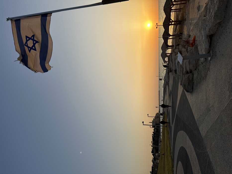 The image depicts a sunset scene on the Tel Aviv promenade, looking south towards Old Jaffa. In the upper right, a worn Israeli flag, light blue and white with a Star of David, flies from a pole against a gradient sky transitioning from clear blue overhead to orange and yellow at the horizon. A faint crescent moon is visible in the upper left sky.
The foreground features a paved promenade with a distinctive dark and light gray wavy pattern. To the left of the promenade, a green grassy area extends. Several tall lampposts line the path. Two individuals are walking away from the camera on the promenade, appearing as dark silhouettes. In the far distance on the left, on an elevated landmass, the buildings of Old Jaffa are visible, including what appears to be the Jaffa Clock Tower and the spire of St. Peter's Church.
To the right of the promenade, a rocky strip separates the path from a sandy beach. On the beach, several open-sided, roofed structures, likely sunshades or pergolas, are partially silhouetted. The Mediterranean Sea stretches to the horizon, with the bright orange sun visible as a distinct disk at sea level. The water reflects the sun's golden light. One or two indistinct figures are present near the beach structures. A small, light-colored sign on a short pole is positioned among the rocks in the lower mid-right.