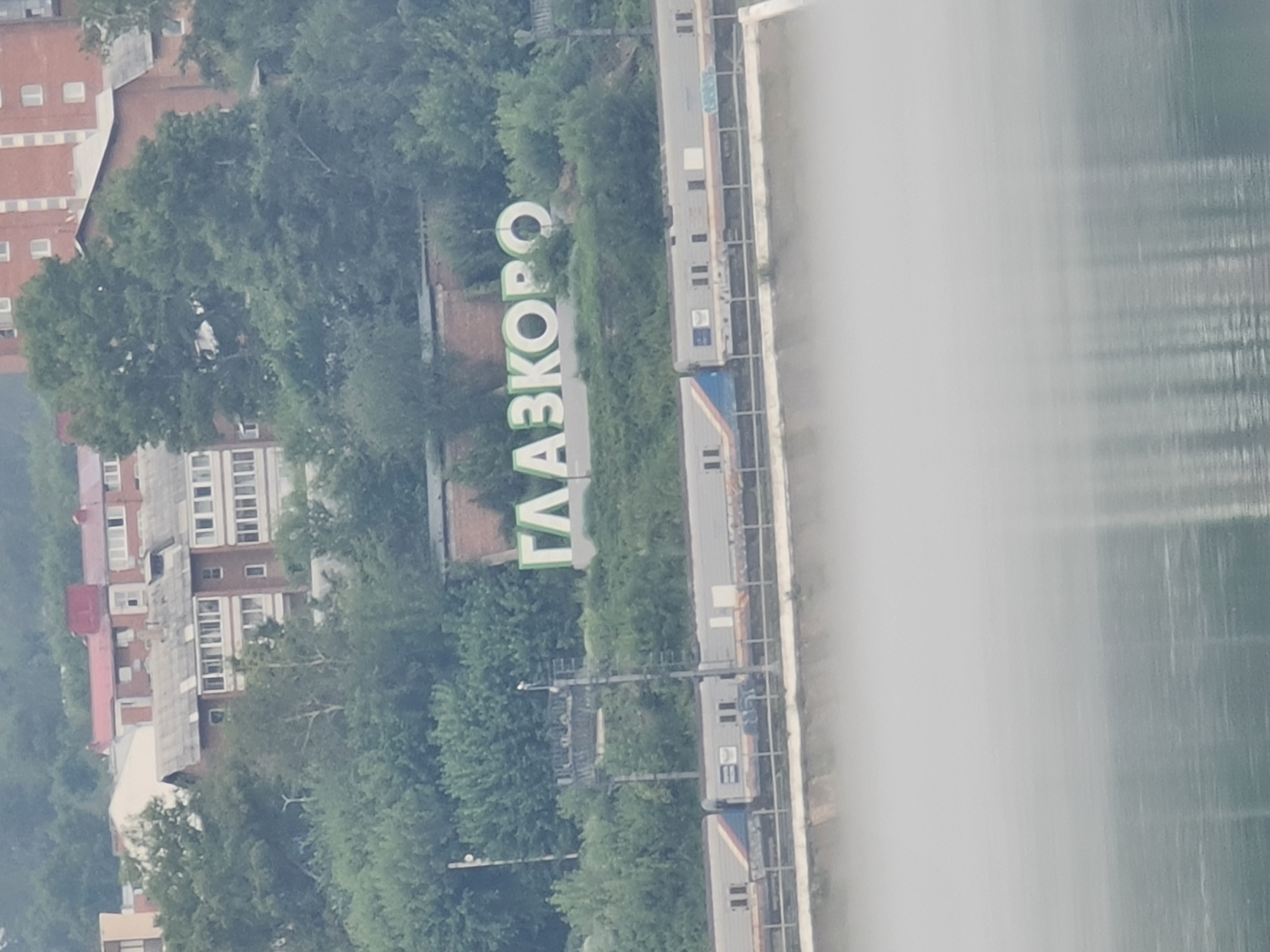 The image shows a section of the city of Glazkovo in Irkutsk, Russia. In the foreground, a train with multiple cars is visible on tracks. Behind the train, large, white, three-dimensional letters spelling "ГЛАЗКОВО" are situated on a grassy slope, forming a landmark. The letters are outlined in green. Above the letters, buildings and trees are visible. The lower portion of the image features a body of water, partially obscured by a layer of mist or fog.