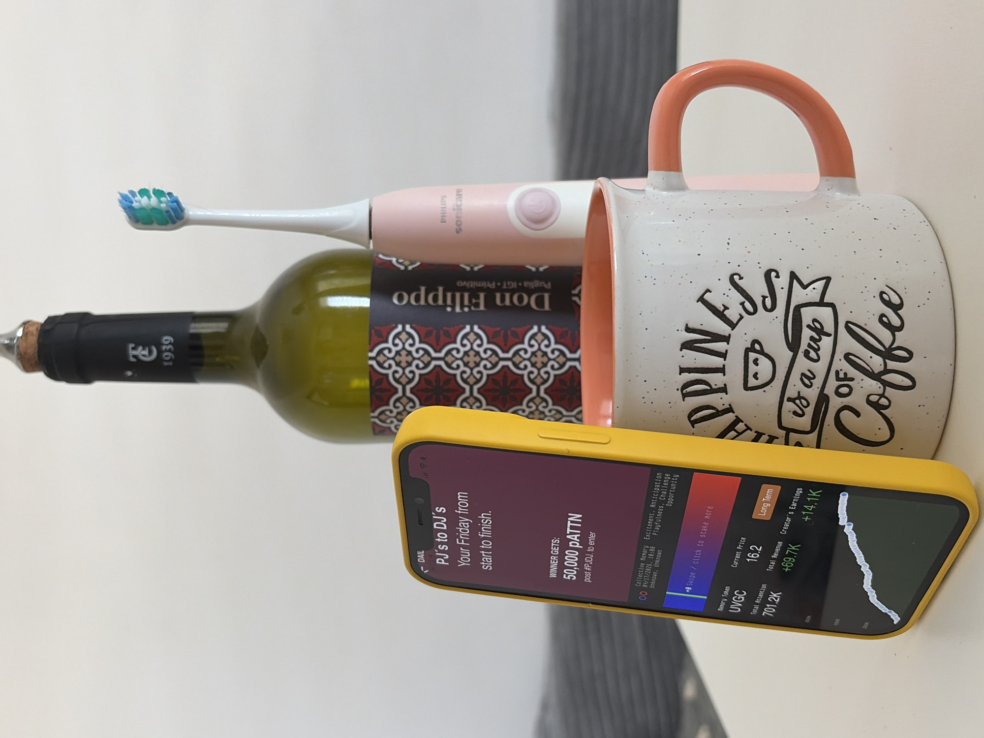 The image displays an arrangement of everyday items lying horizontally on a light-colored surface, likely a table or counter. The objects, from back to front, include a green wine bottle, a pink and white electric toothbrush, a white and orange coffee mug, and a smartphone in a yellow case.
The wine bottle has a black label featuring "Don Filippo" and "Puglia IGT Primitivo," with the year "1939" also visible. It is topped with a cork. Resting partially on the wine bottle's label is a pink Philips Sonicare electric toothbrush, showing its blue and green bristles and the Philips Sonicare branding. To the right, a white speckled coffee mug with an orange handle is visible. Its side is adorned with text, part of which reads, "HAPPINESS is a cup Coffee."
In the foreground, angled towards the viewer, is a smartphone in a bright yellow protective case. Its screen is illuminated, displaying what appear to be two different application interfaces. One section, labeled "DAIL," features "PJ's to DJ's" and the text "Your Friday from start to finish." It advertises a contest: "WINNER GETS: 50,000 PATTN" and instructs to "post #PJDJ. to enter." This app also lists keywords such as "Collective Memory," "Excitement, Anticipation," "Playfulness, Challenge," and "Opportunity," with a date "04/17/2026, 18:00." The other section of the screen shows financial or crypto-related data under "Memory Token UVGC," with a "Current Price 16.2," "Total Attention 701.2K," "Total Revenue +69.7K," and "Creator's Earnings +14.1K," accompanied by a graph showing a positive, upward trend over the "Long Term."
The scene captures a blend of personal care, leisure, and digital activity, set in what is understood to be Budva, Montenegro.