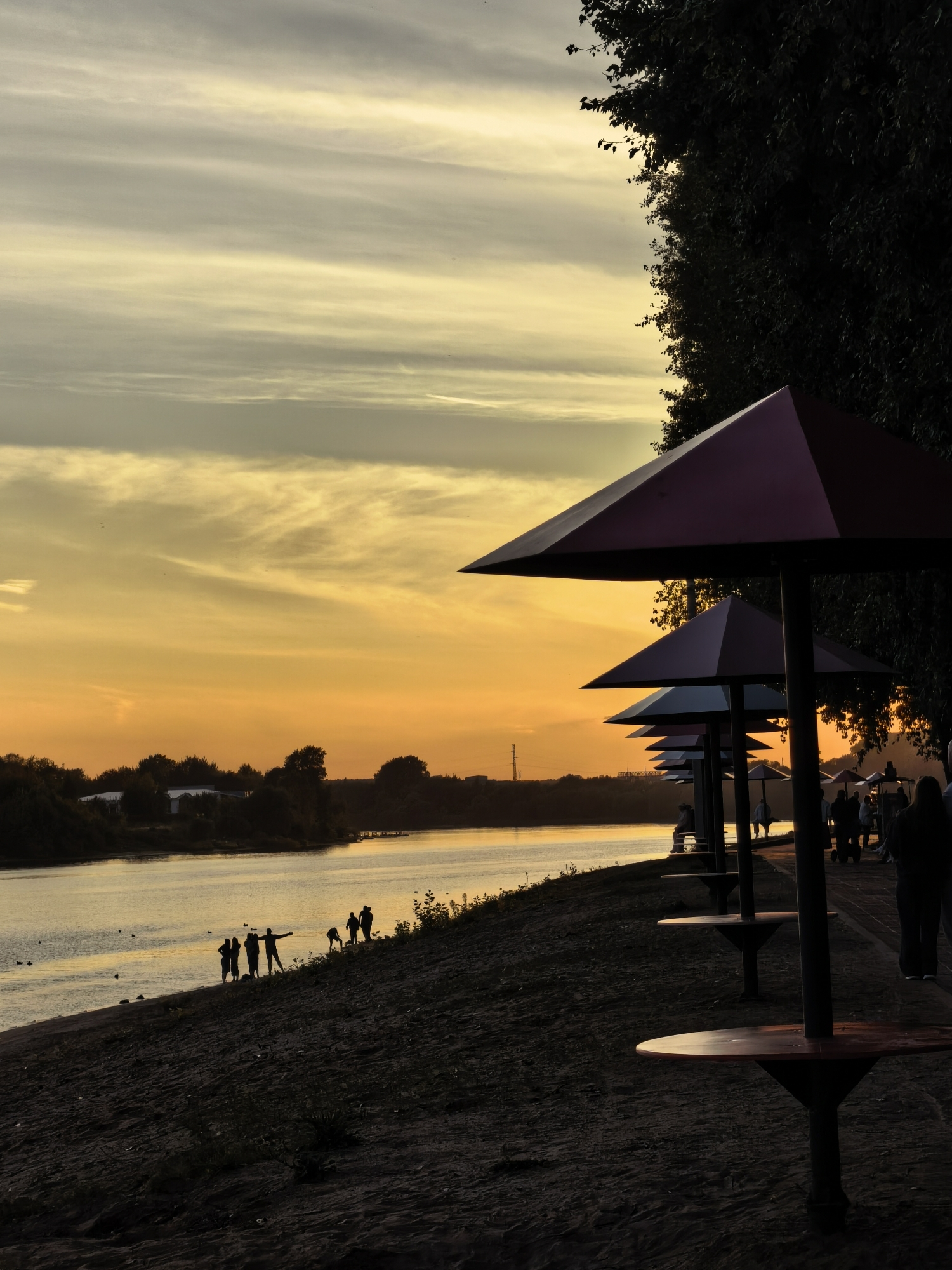The image depicts a sunset scene on the bank of the Volga River in Tver, Russia. In the foreground, several outdoor umbrella tables, set up for seating, line a sandy embankment. Beyond the embankment, the calm waters of the Volga reflect the warm hues of the setting sun. A group of silhouetted figures are gathered near the water's edge, some appearing to be playing or interacting. Across the river, the opposite bank is lined with trees and vegetation, with hints of buildings in the distance. The sky is filled with soft, wispy clouds illuminated by the golden and orange glow of the sunset. A tall transmission tower is visible on the far bank, adding a touch of industrial presence to the otherwise natural landscape. The overall atmosphere is serene and tranquil, capturing the beauty of a late summer evening.