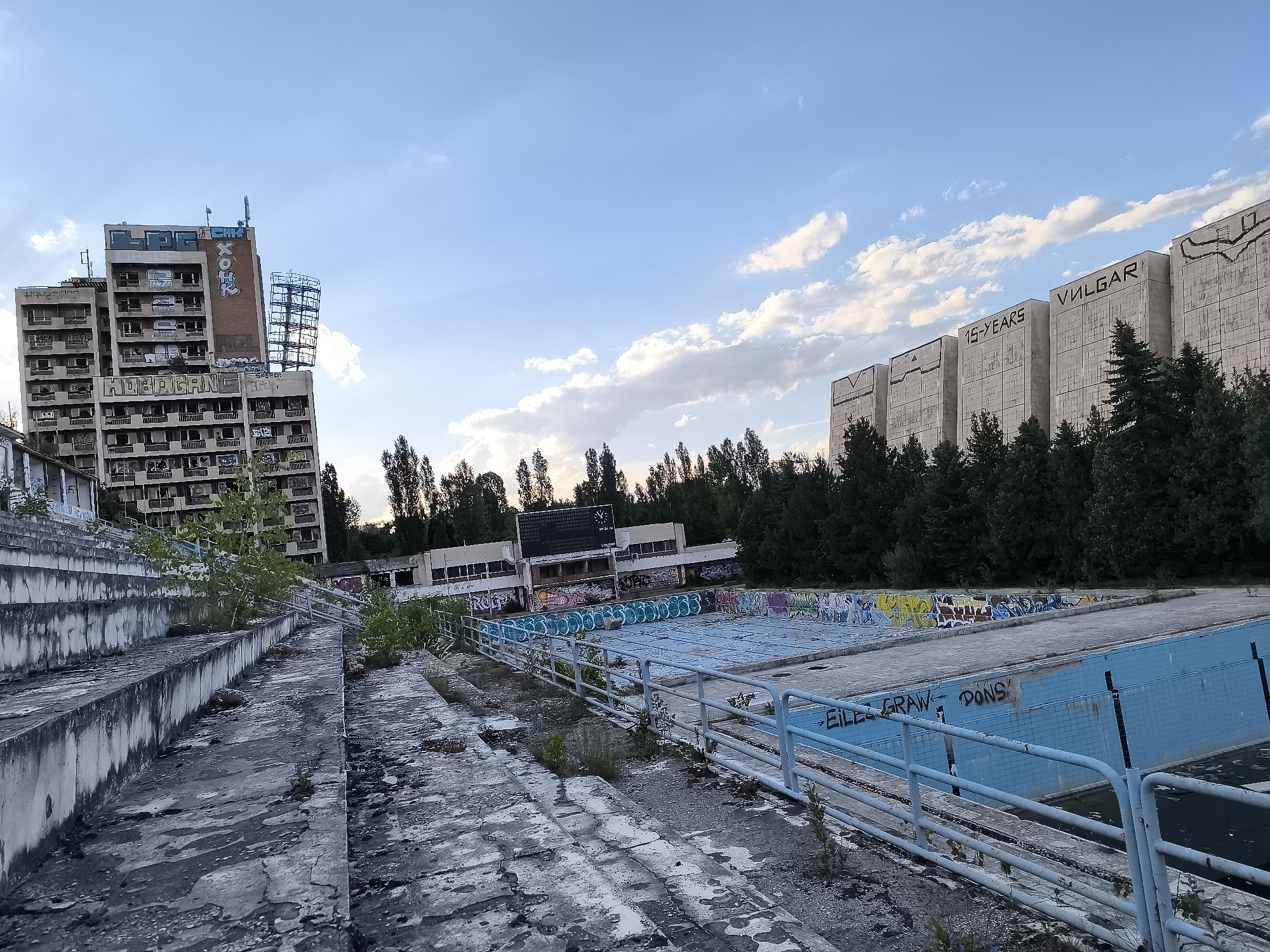 The image depicts an abandoned outdoor swimming pool complex in Sofia, Bulgaria, with dilapidated buildings and graffiti-covered structures in the background. In the foreground, concrete bleachers are weathered and overgrown with weeds. A light blue metal railing separates the bleachers from the empty pool area. The pool itself is empty, with bright blue tiles lining its bottom and sides, some of which are cracked or missing. The pool area is heavily adorned with colorful graffiti, as are the surrounding concrete surfaces.
In the mid-ground, a long, low building with broken windows and more graffiti stretches along the side of the pool. Behind this building, a dense line of trees separates the pool complex from the background. To the left of the pool complex, a tall, multi-story building stands abandoned, with numerous windows and balconies, also covered in graffiti. A metal scaffolding structure is attached to the side of this building. To the far right of the image, several large, Brutalist-style concrete buildings are visible, also featuring some graffiti and inscriptions, including "VULGAR" and "15-YEARS". The sky above is a pale blue with scattered white clouds. The overall scene conveys a sense of decay and abandonment in an urban environment.